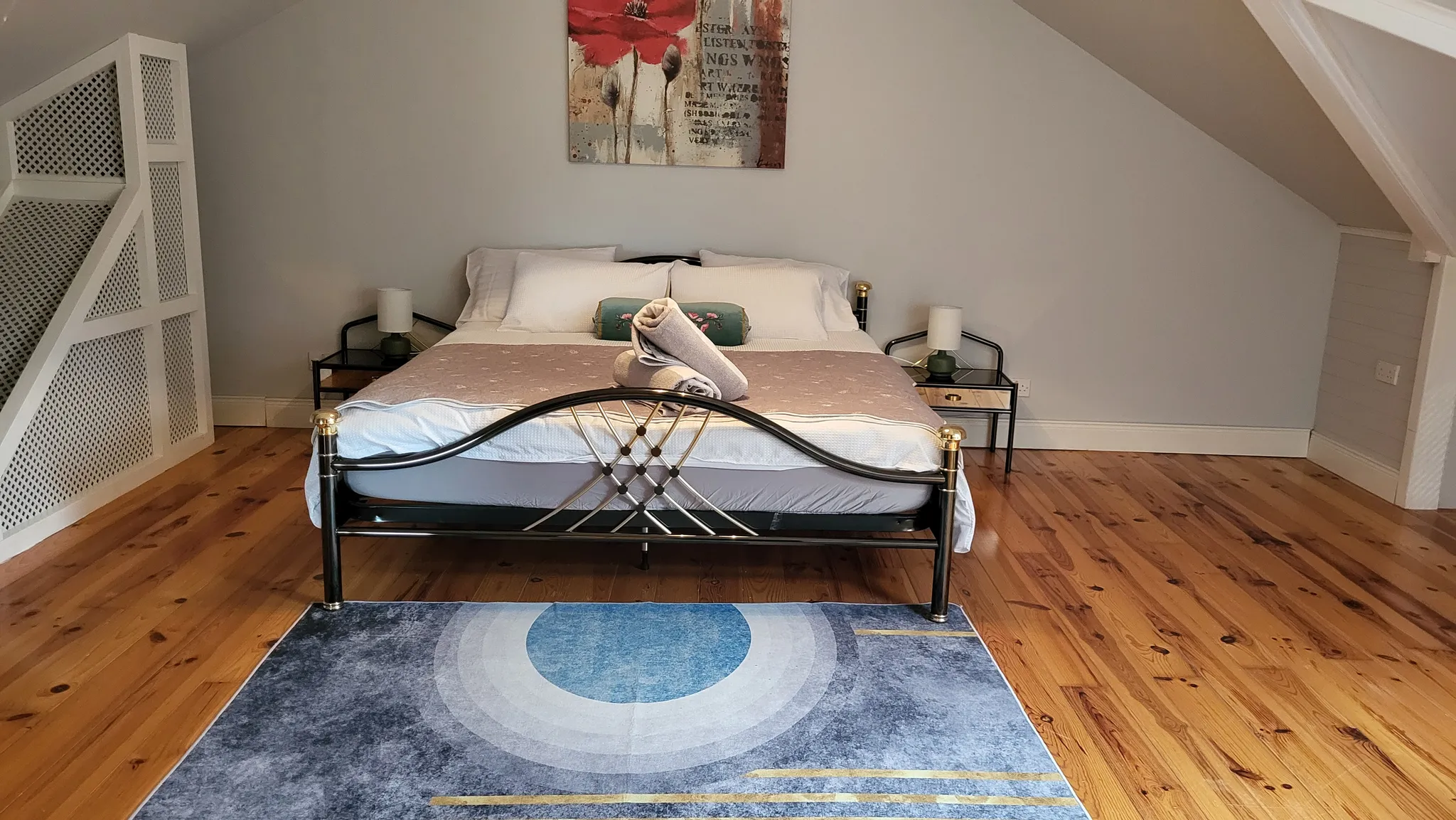 Bedroom with a double bed centered under a sloped ceiling, two bedside tables with lamps, folded towels on the bed, a decorative wall painting above the headboard, and a rug on a wooden floor.