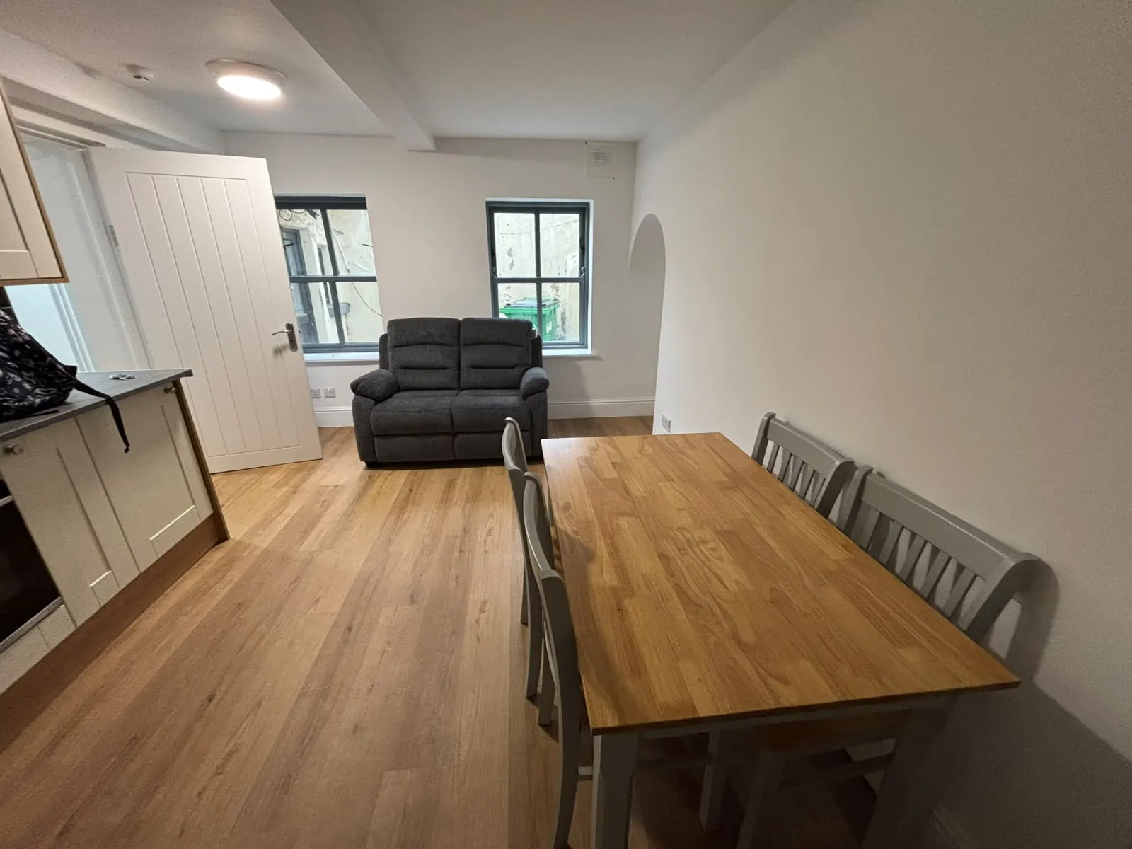Combined living and dining area with a wooden dining table and chairs, a two-seat grey sofa, two windows, and a partially open door near a kitchen counter.