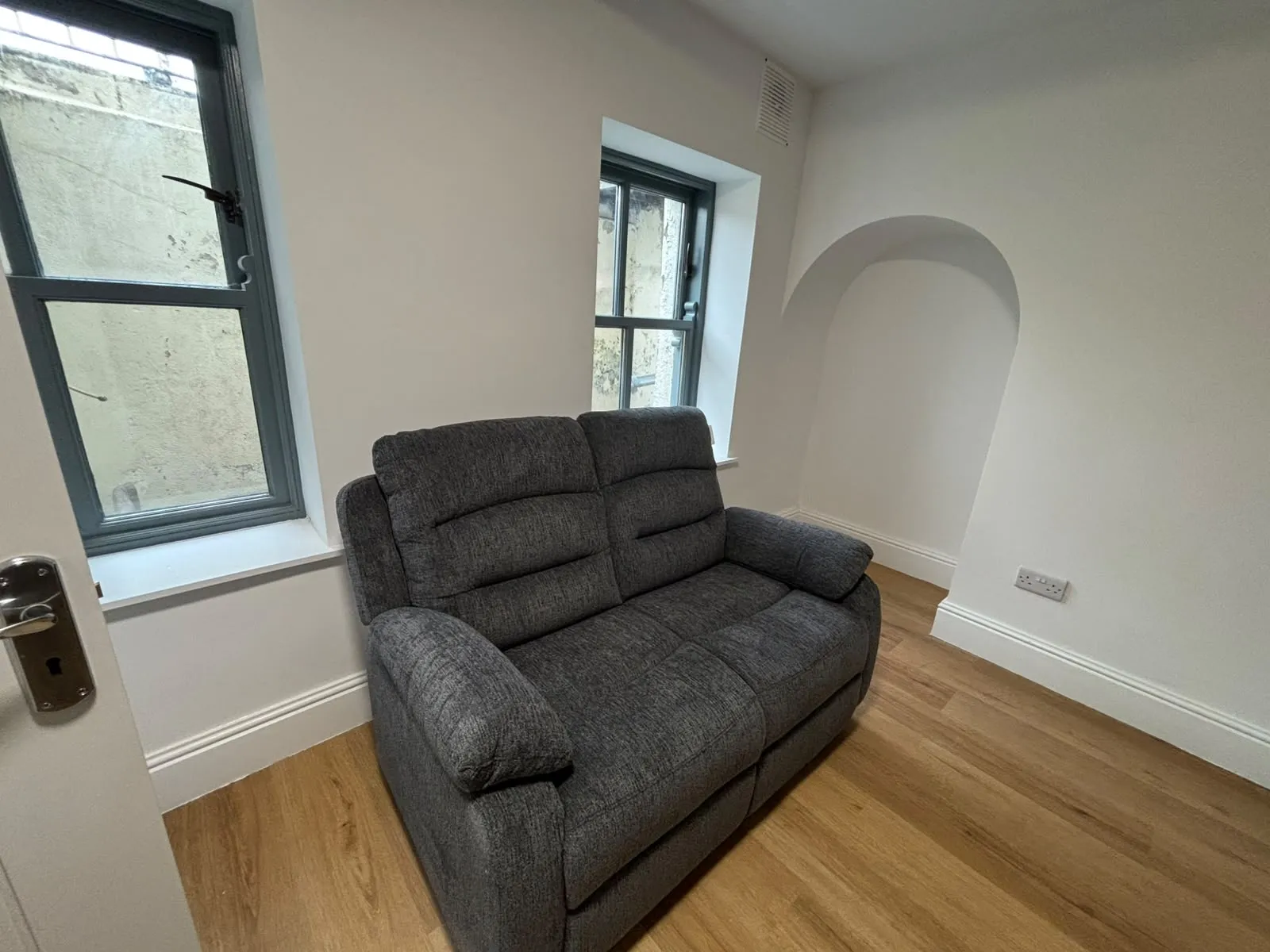 Living room with a two-seater gray upholstered sofa, two windows with gray frames, an arched wall niche, and wood-effect flooring.