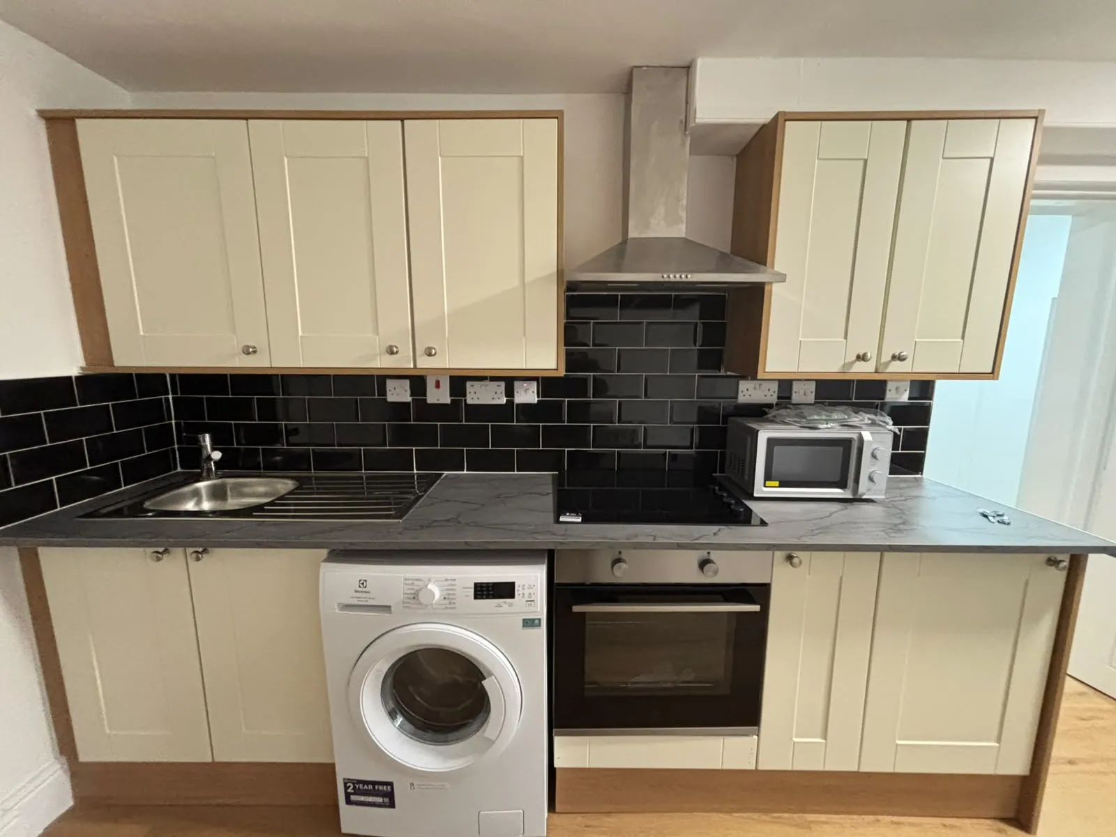 Small kitchen with cream cabinets, black subway tile backsplash, stainless sink, countertop hob and oven, washing machine and microwave