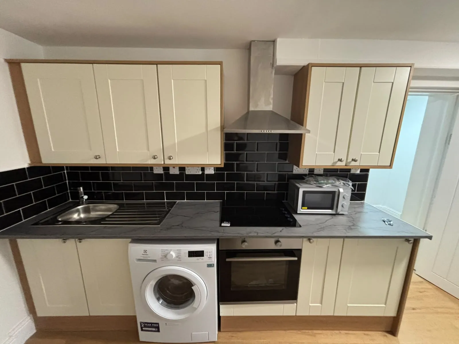 Kitchen with upper and lower cabinets, stainless steel sink, black tiled backsplash, electric hob with extractor hood, oven, washing machine and countertop microwave.