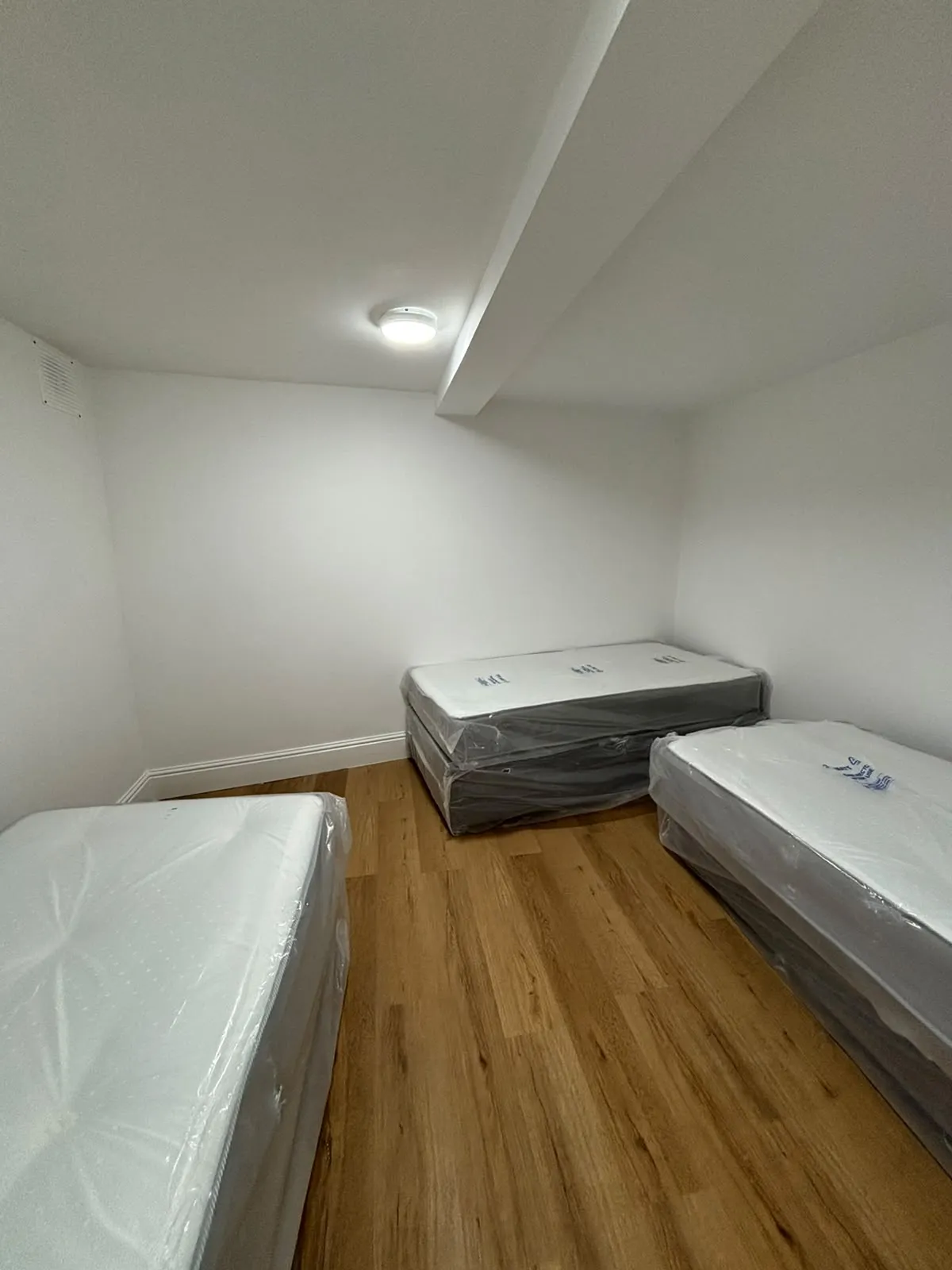 Small bedroom with three single mattresses still wrapped in plastic on a wood floor and a ceiling light
