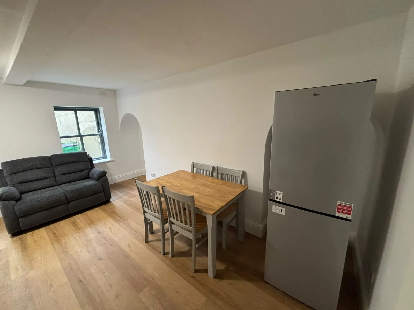Living area with a gray two-seat sofa, a wooden dining table with four chairs, a tall silver refrigerator, and a window in a white-walled room.