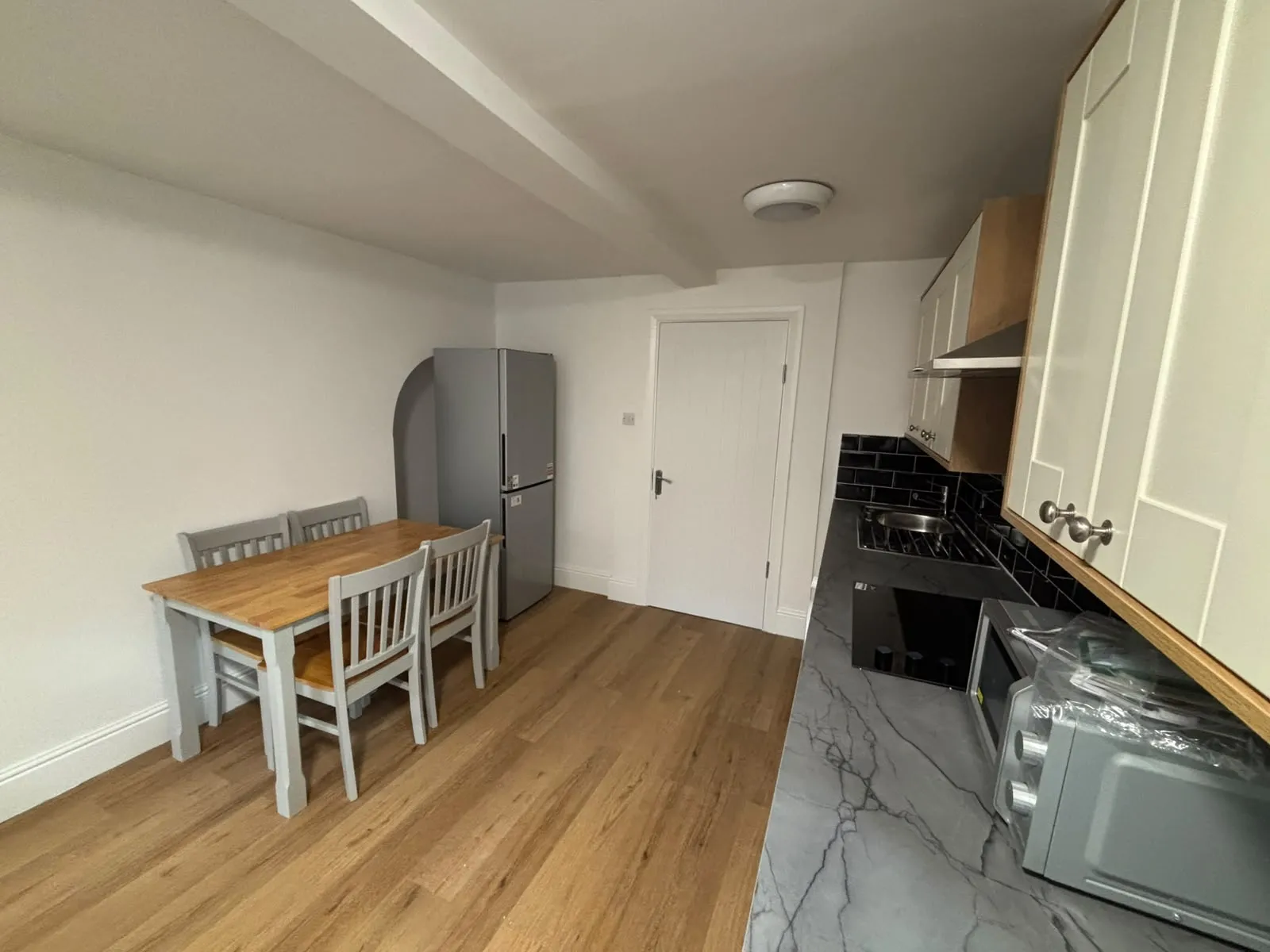 Kitchen with a wooden dining table and four chairs, stainless refrigerator against a white wall, and a countertop with sink, stovetop and microwave.