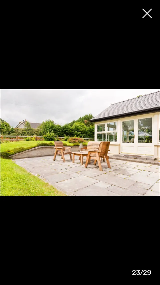 Exterior patio area with wooden furniture, stone paving, and manicured garden. Modern house with large windows and grey roof visible in background.