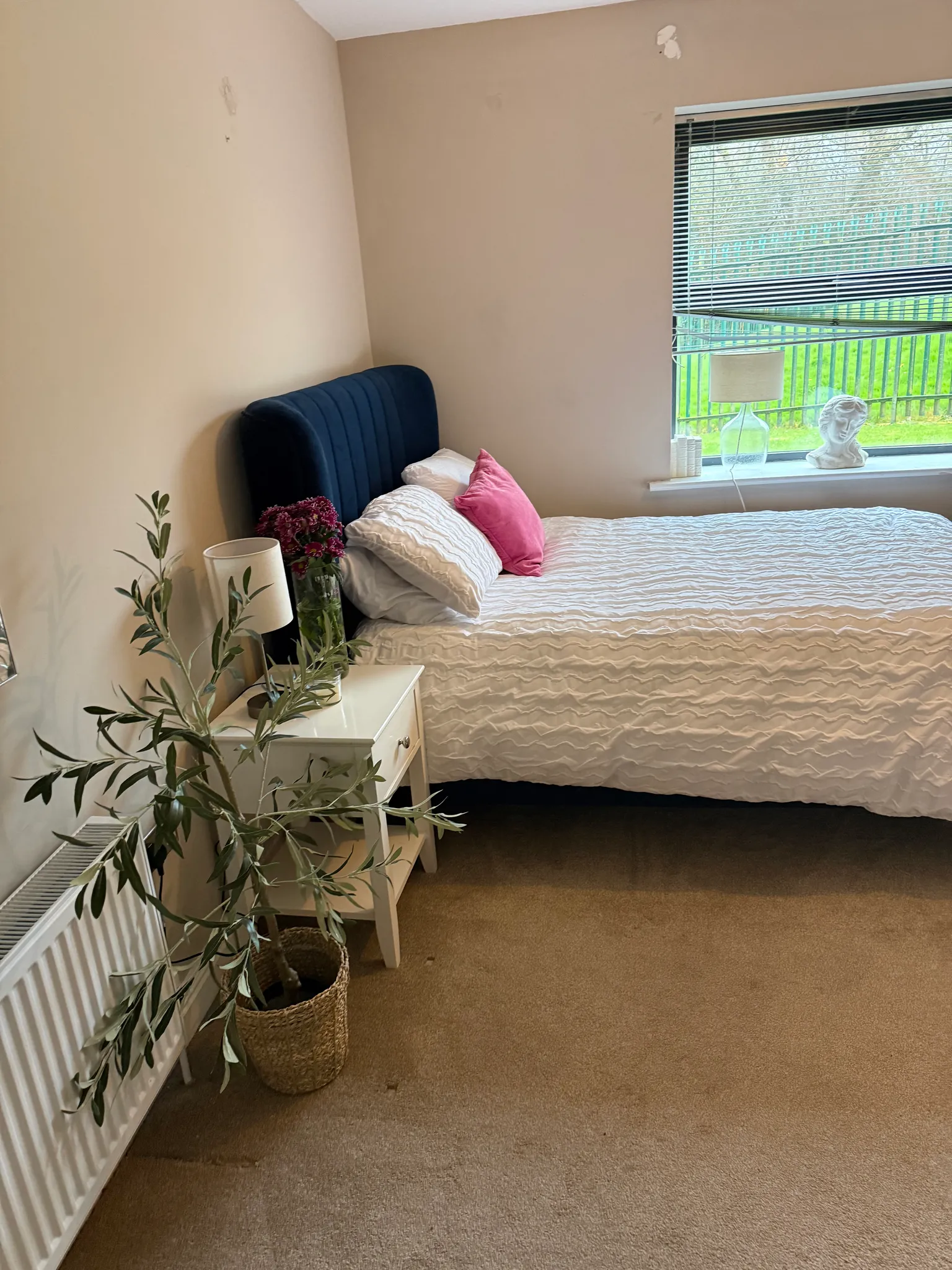 Bedroom with a bed, dark upholstered headboard, bedside table with lamp and flowers, potted plant, and a window with horizontal blinds