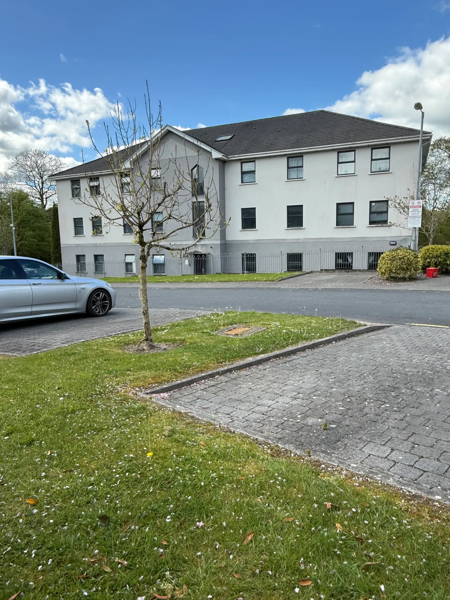 Exterior view of a three-story apartment building seen from the street with grass and a small tree in the foreground and a parked car at left.