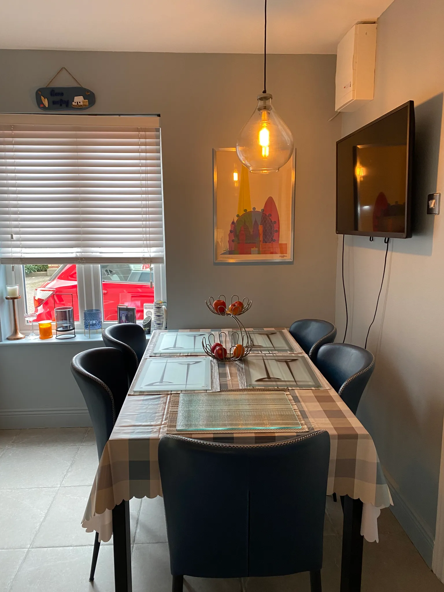 Dining room with a rectangular table set for four, pendant light above, window with closed blinds, wall-mounted TV and framed artwork.