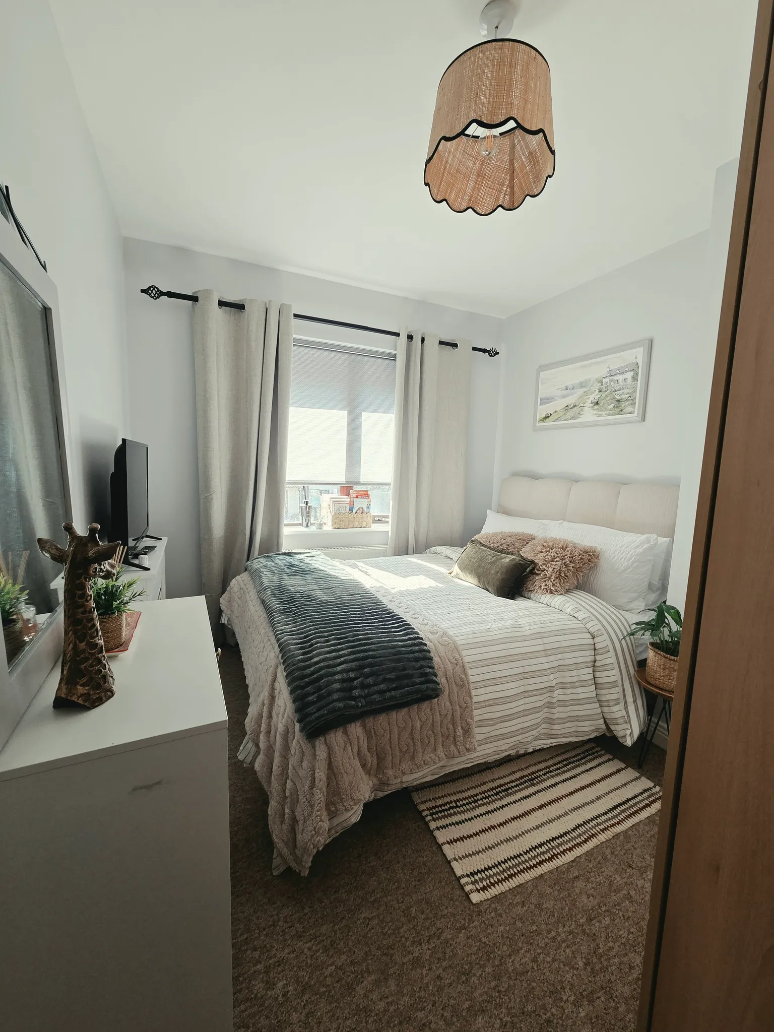 Bedroom with a double bed, window with curtains, dresser and TV, ceiling light and bedside pillows.