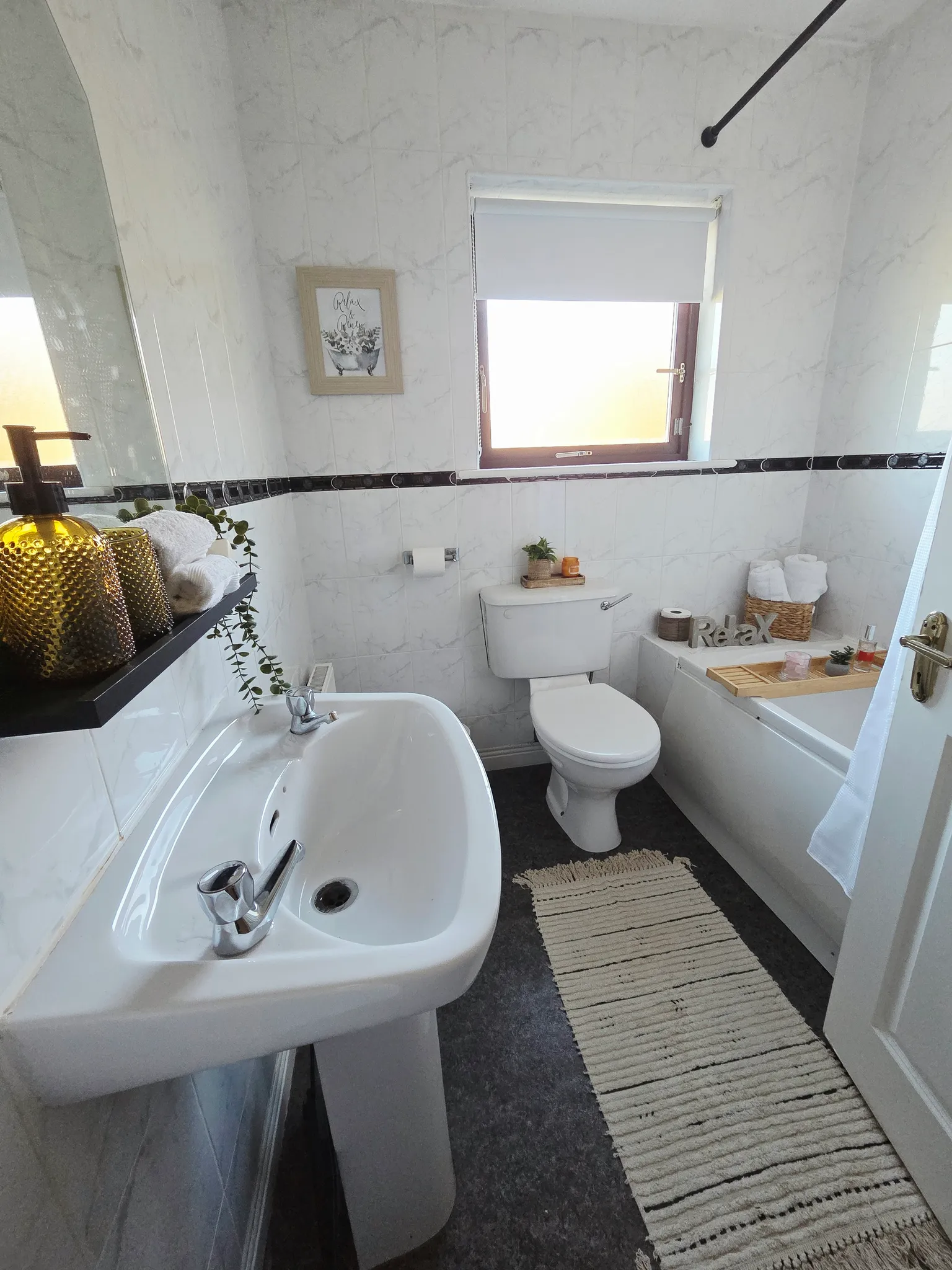 Bathroom with a sink and mirror on the left, a toilet below a window with a blind, and a bathtub with a wooden tray on the right.