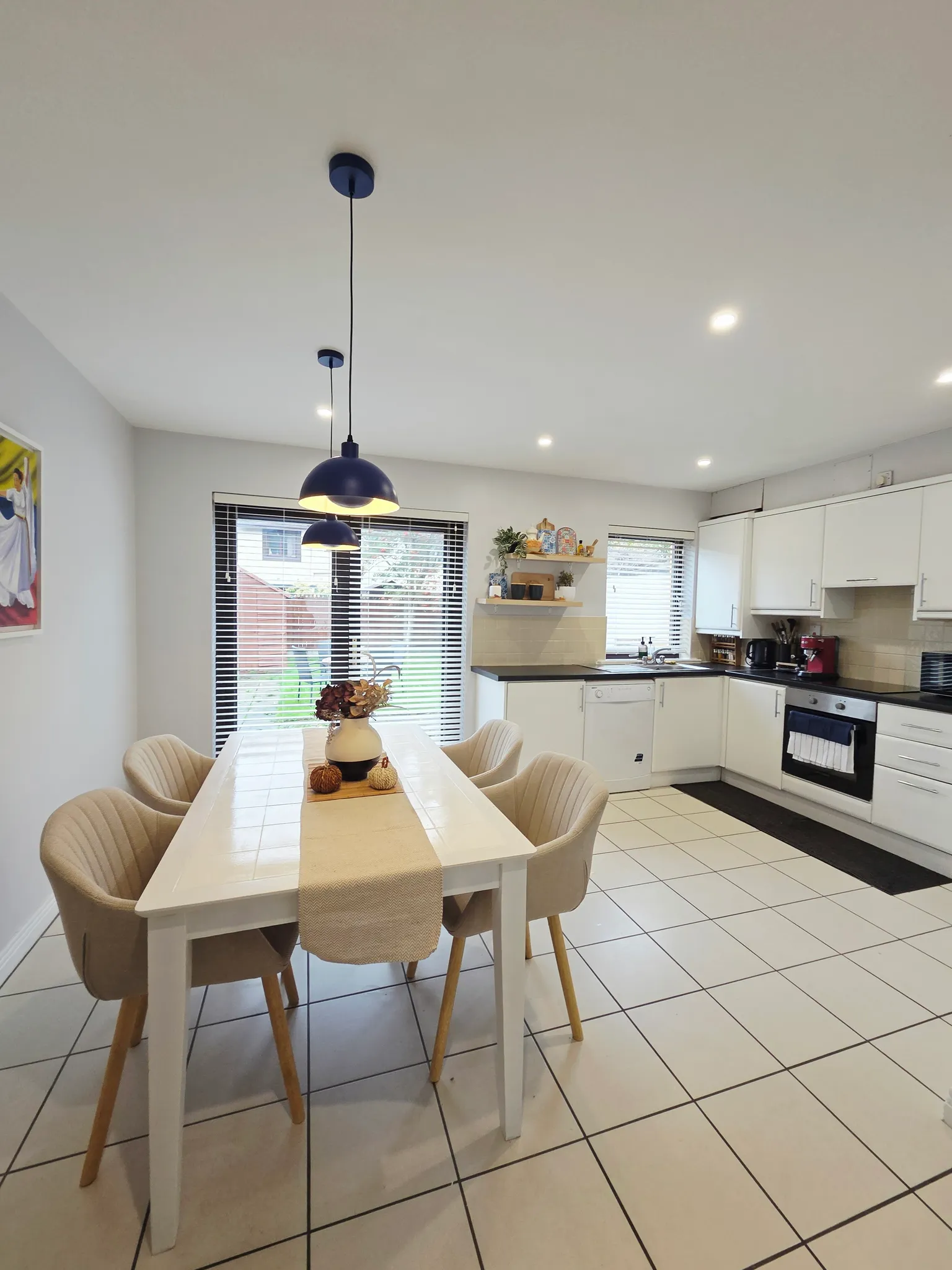 Open-plan dining area with a rectangular white table, four upholstered chairs, two pendant lights above the table and a kitchen with white cabinets and appliances in the background