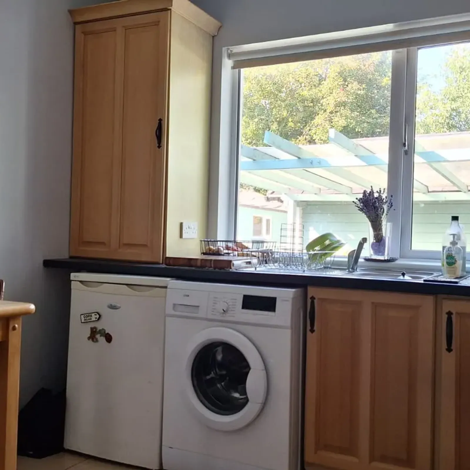 Kitchen with wooden cabinets, a countertop under a large window, a front-loading washing machine, a small fridge, kettle, and a wooden breakfast table and chair.