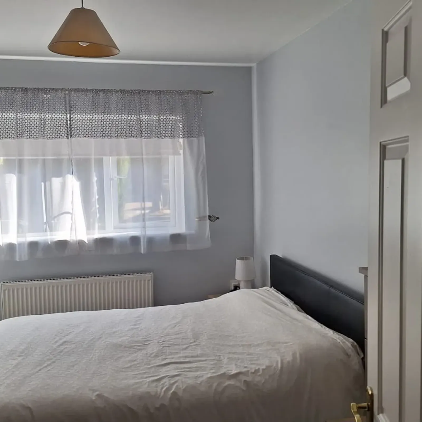 Bedroom with a bed, window with sheer curtains, radiator below the window, bedside lamp and pendant light