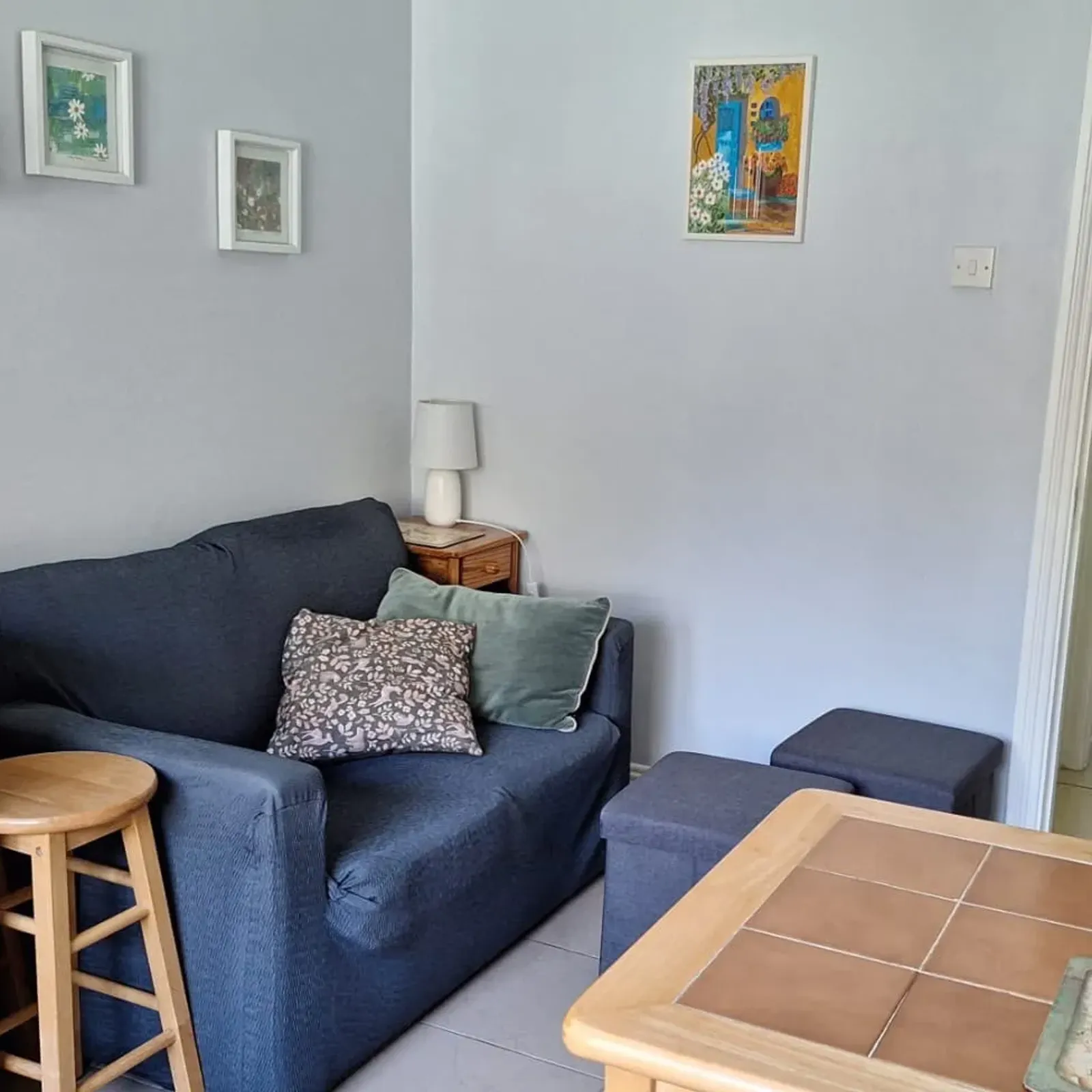 Small living room area with a dark two-seat sofa, two throw pillows, a side table with a lamp, two ottomans, a wooden counter with stools, and a doorway leading to a bedroom.