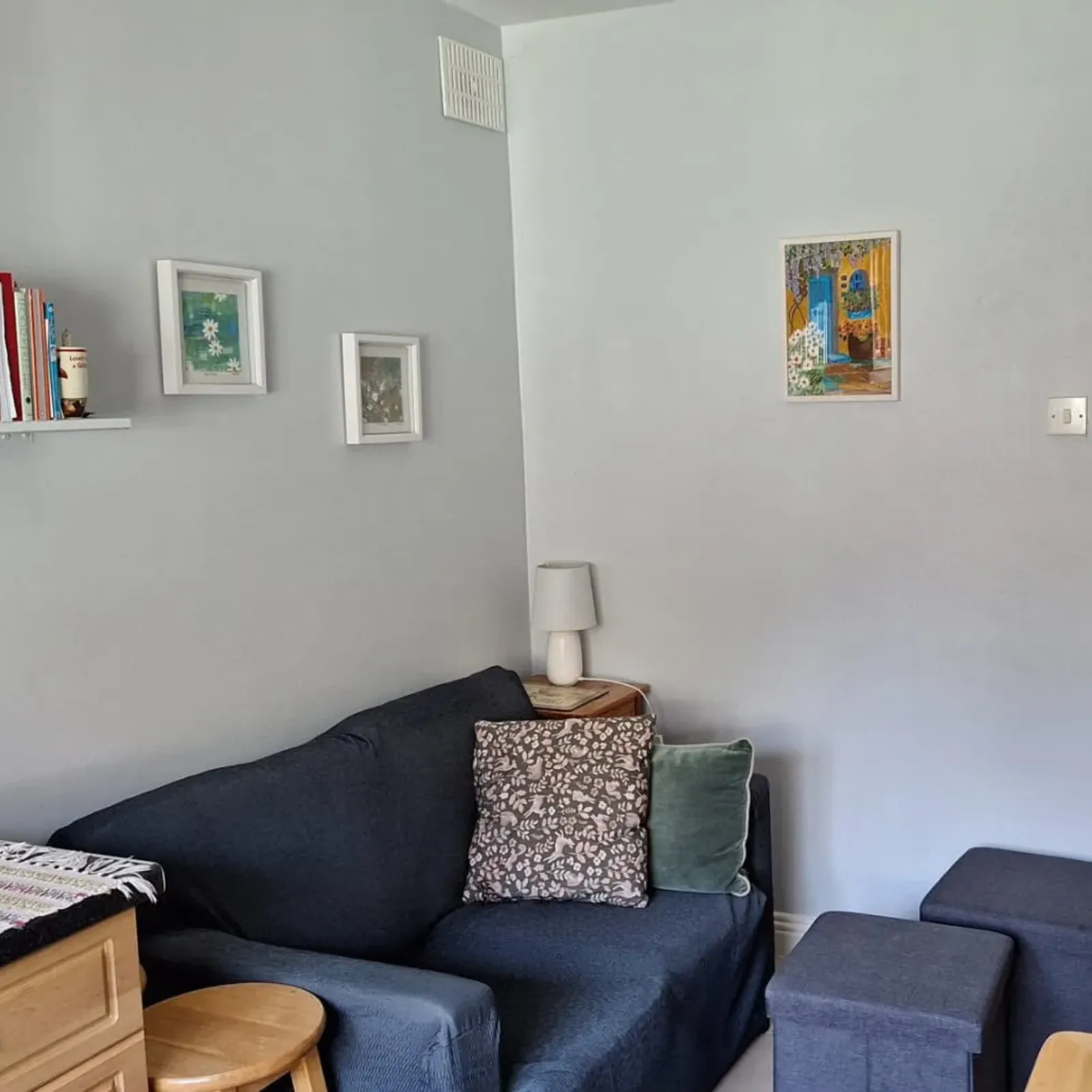 Small living room with a dark two-seat sofa, two patterned cushions, a side table with a lamp, a wall shelf of books and framed artworks, and two square ottomans near a doorway.