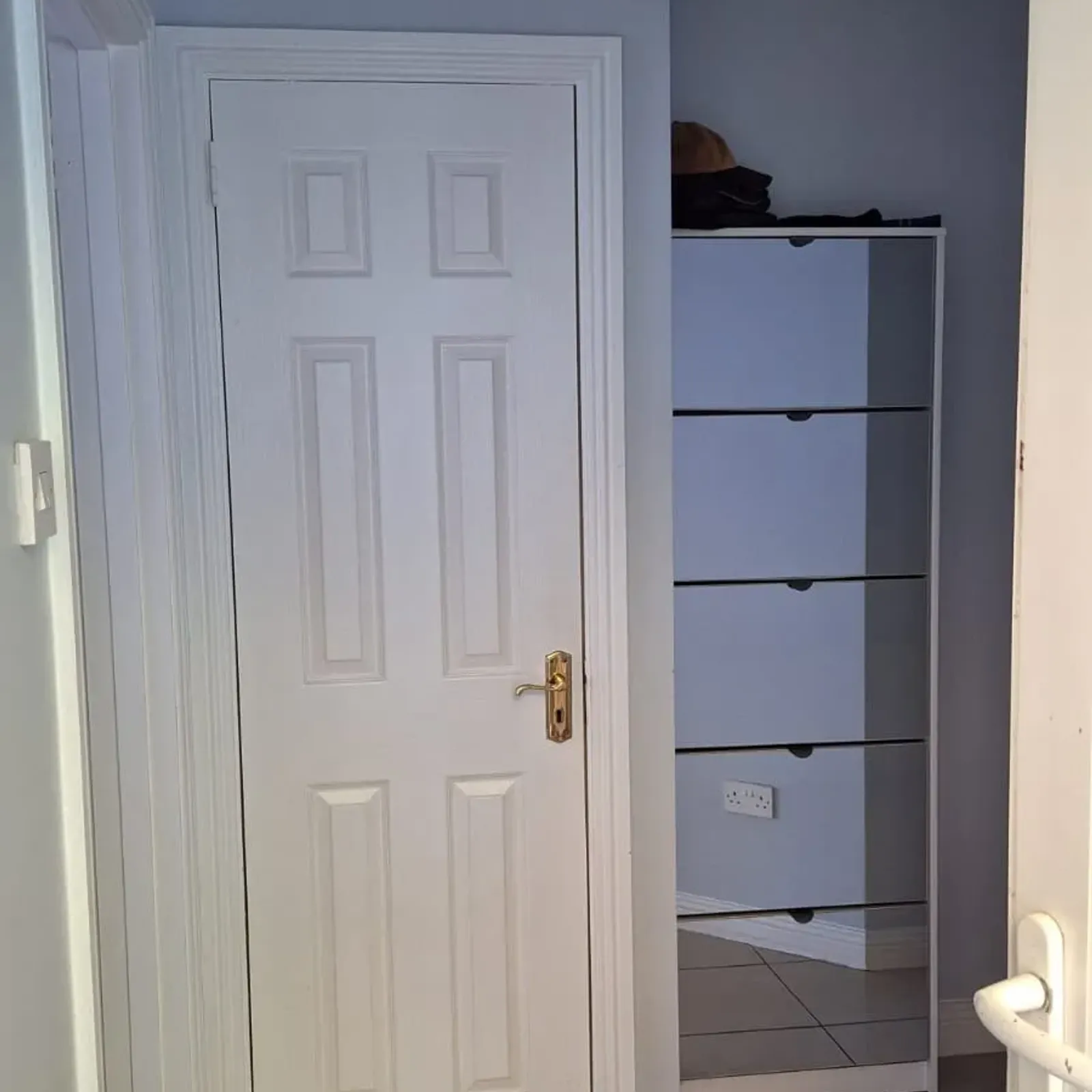 Hallway with a closed white paneled door, tiled floor, radiator at left and stacked storage shelves visible in a recessed alcove to the right.