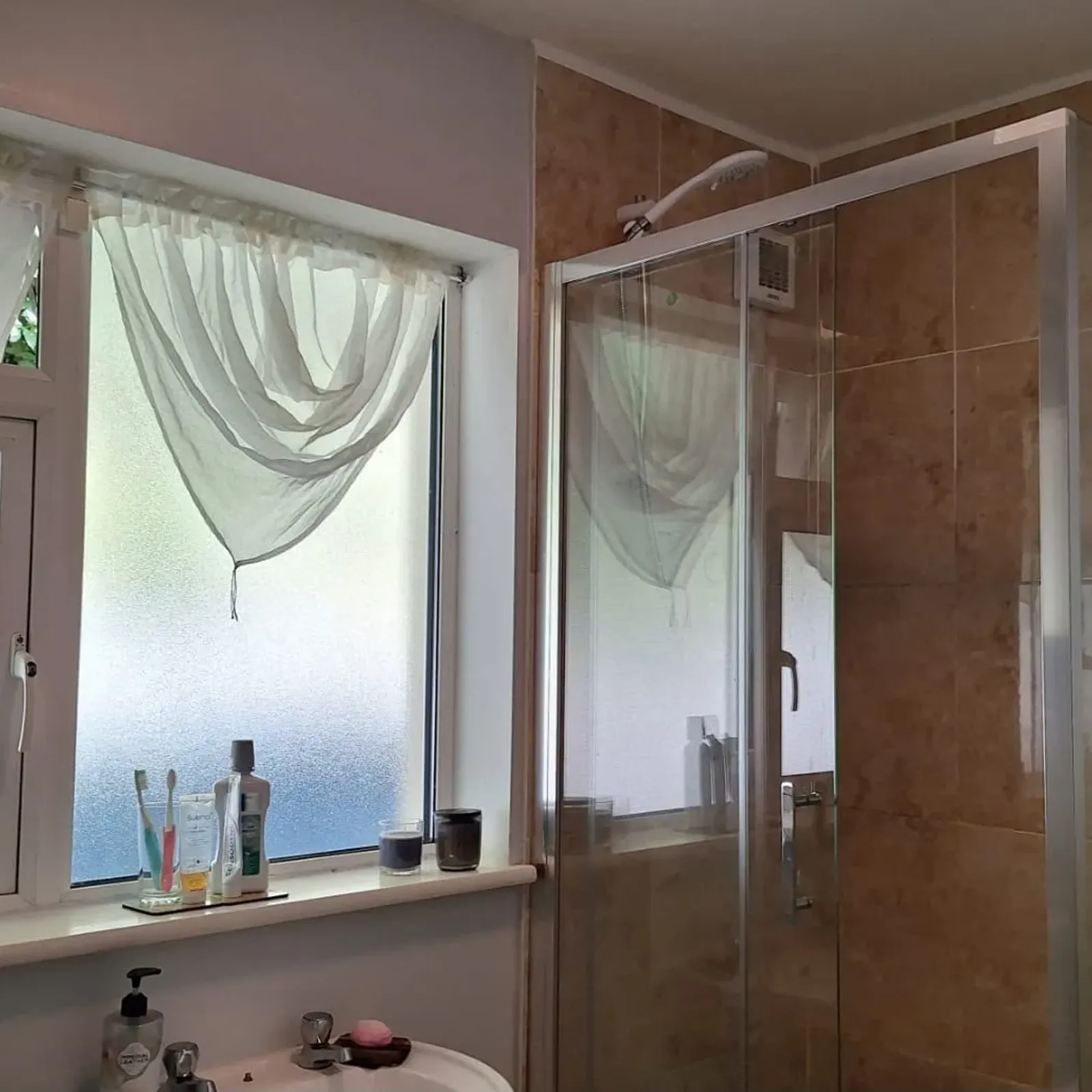 Bathroom with a frosted window and sheer curtain, sink with toiletries on the sill, and a glass-enclosed tiled shower with a showerhead and caddy.