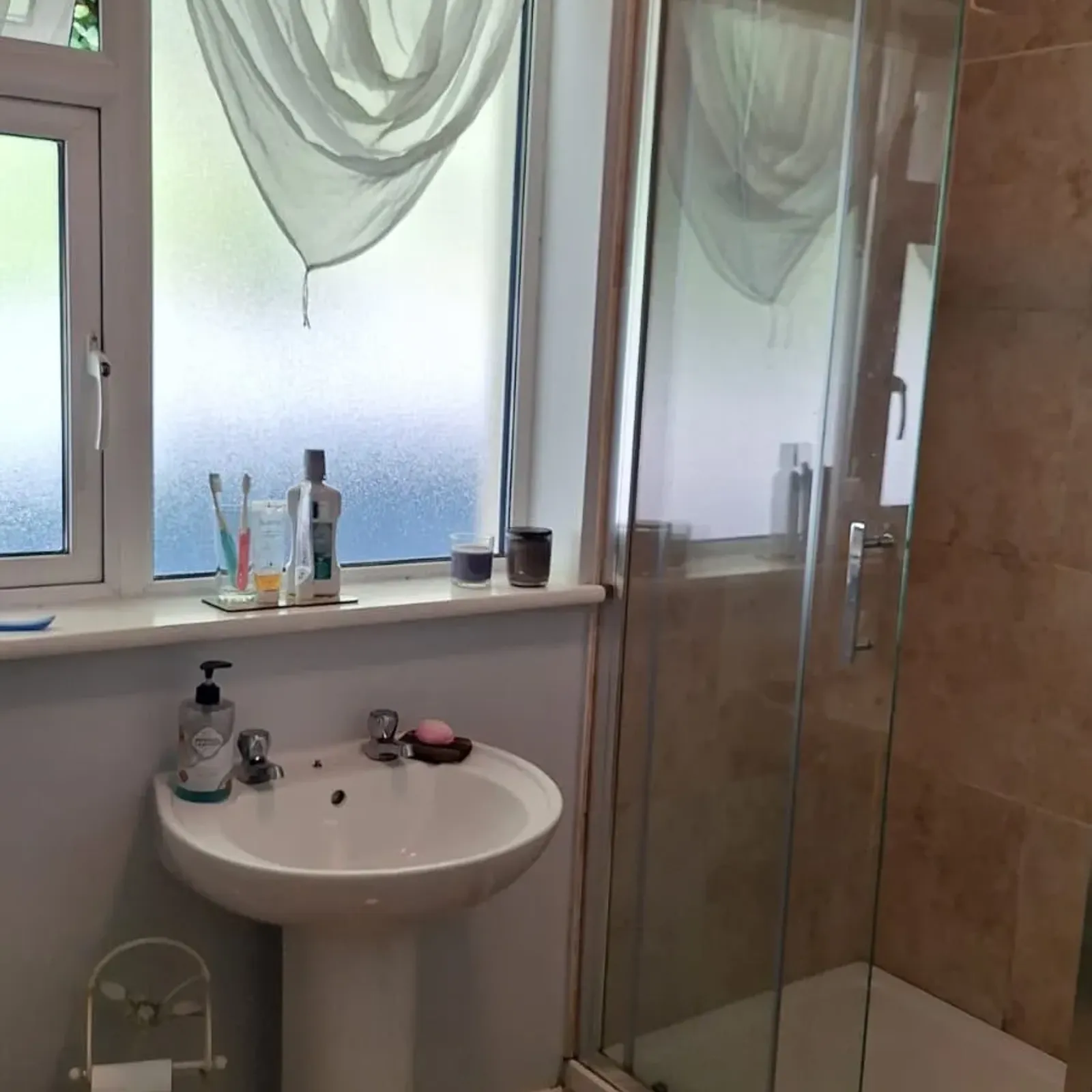 Bathroom with a pedestal sink, frosted window with sheer curtain, glass-enclosed shower, and toiletries on the windowsill.