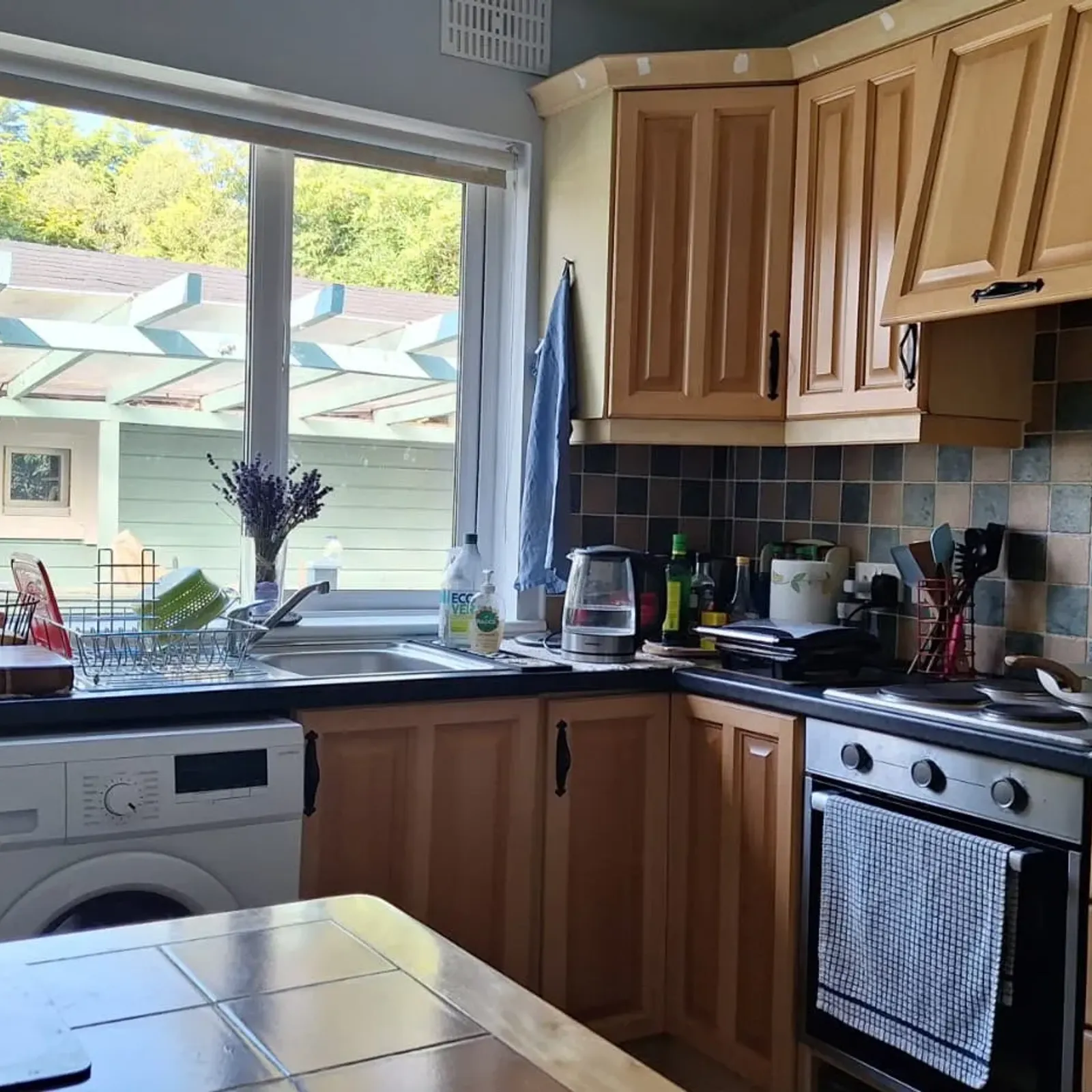 Kitchen with wooden cabinets, tiled backsplash, countertop with sink and kettle, oven and washing machine under the counter, and a large window above the sink.