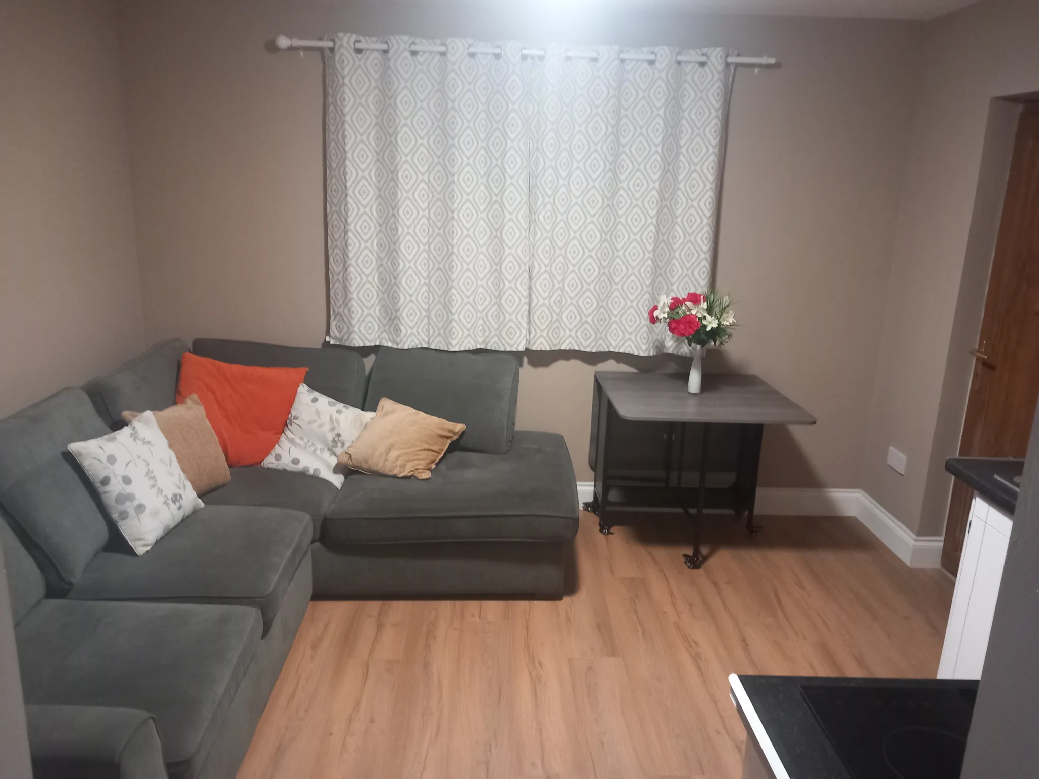 Living room with gray sectional sofa, decorative pillows, wooden side table with potted flowers, patterned curtains, and wood flooring