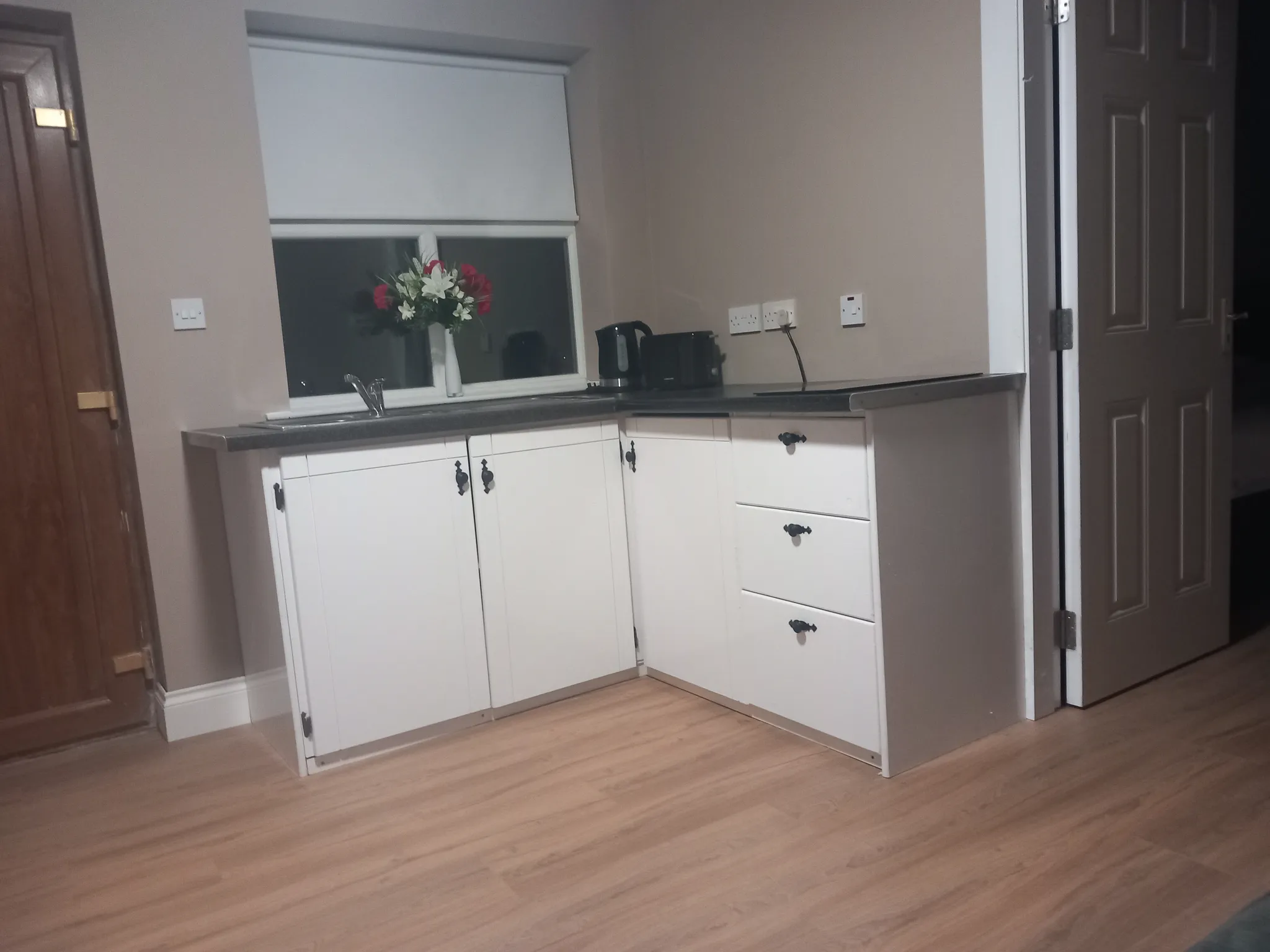 Kitchen with white cabinetry, dark countertop, window with blinds, vase of flowers, and wooden flooring