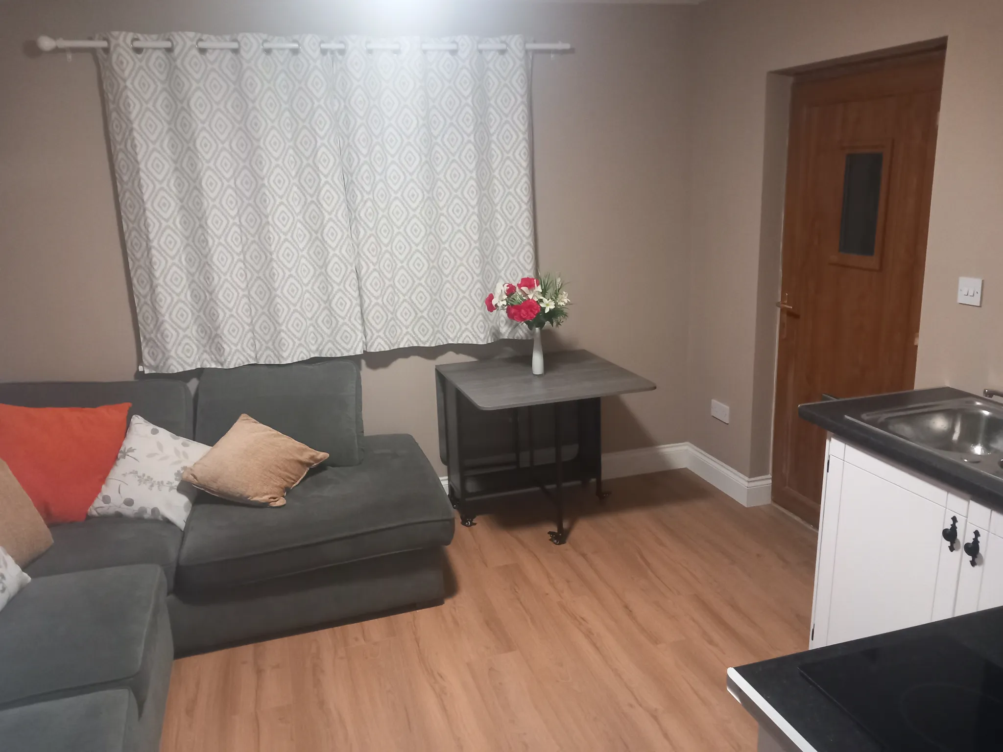 Open-plan living and kitchen area with gray sofa, patterned curtains, wooden side table with flowers, wood flooring, and white kitchen cabinetry with stovetop visible on the right