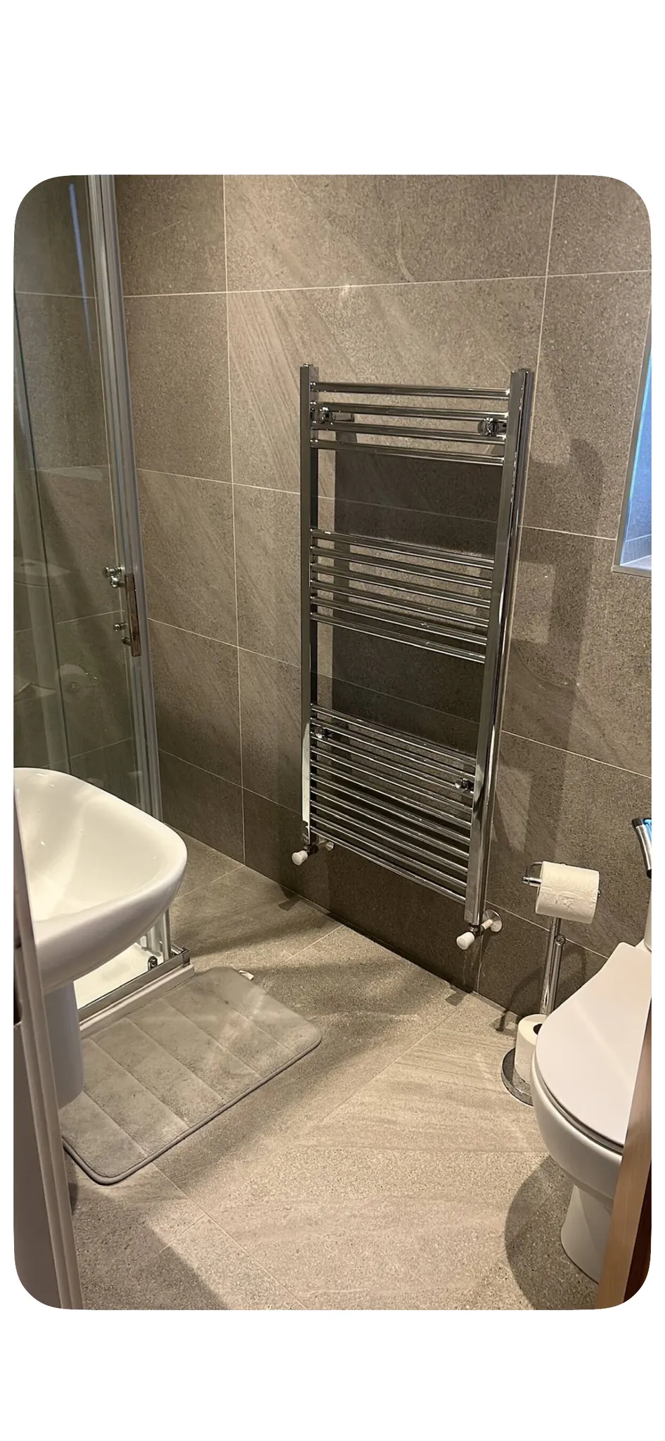 Bathroom with wall-mounted chrome towel radiator, pedestal sink, glass shower door, toilet, toilet paper holder and floor mat.