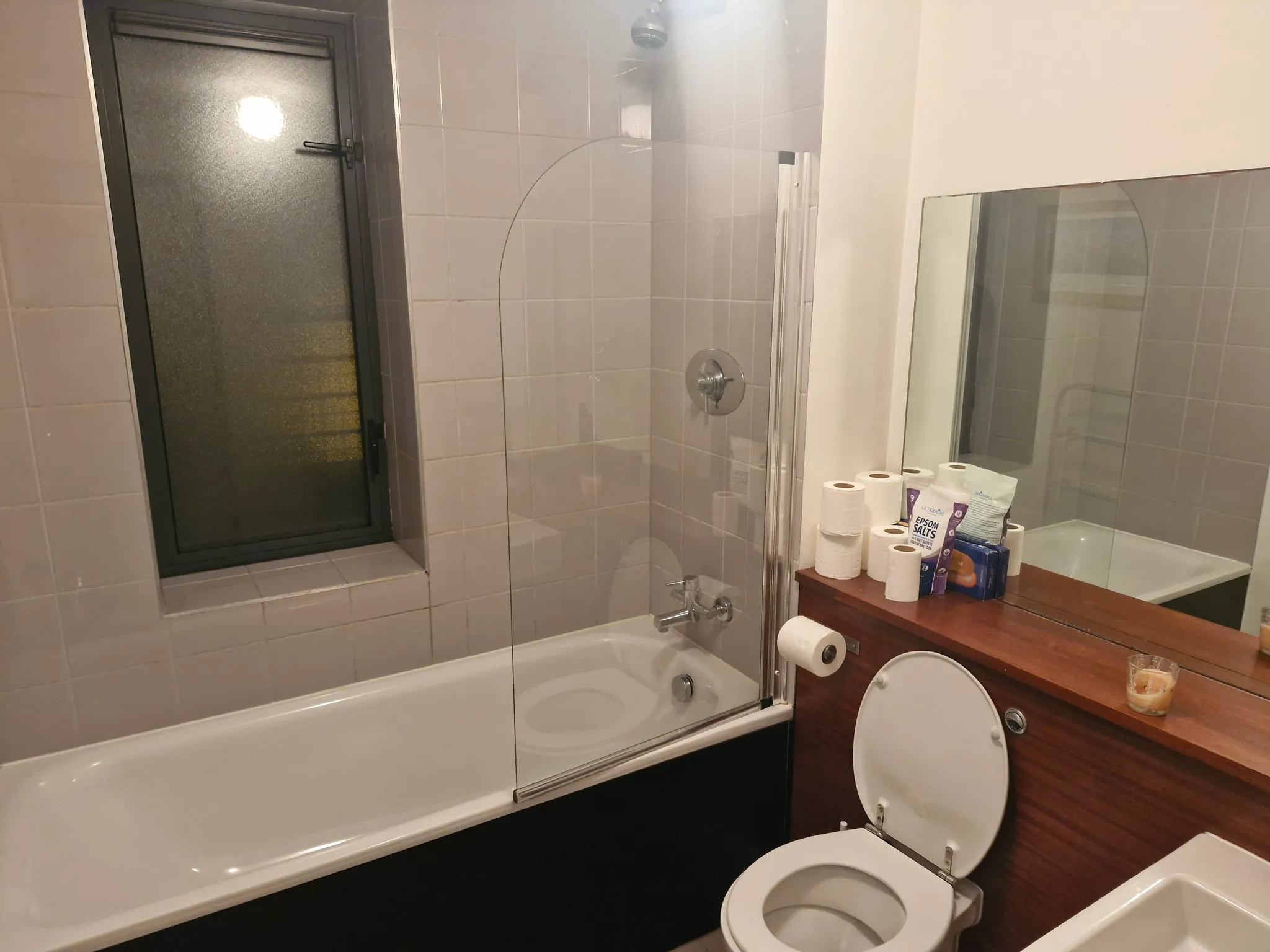 Bathroom with a bathtub and glass shower screen, tiled walls, frosted window, toilet with open seat, countertop with mirror and stacked toilet paper and toiletries.