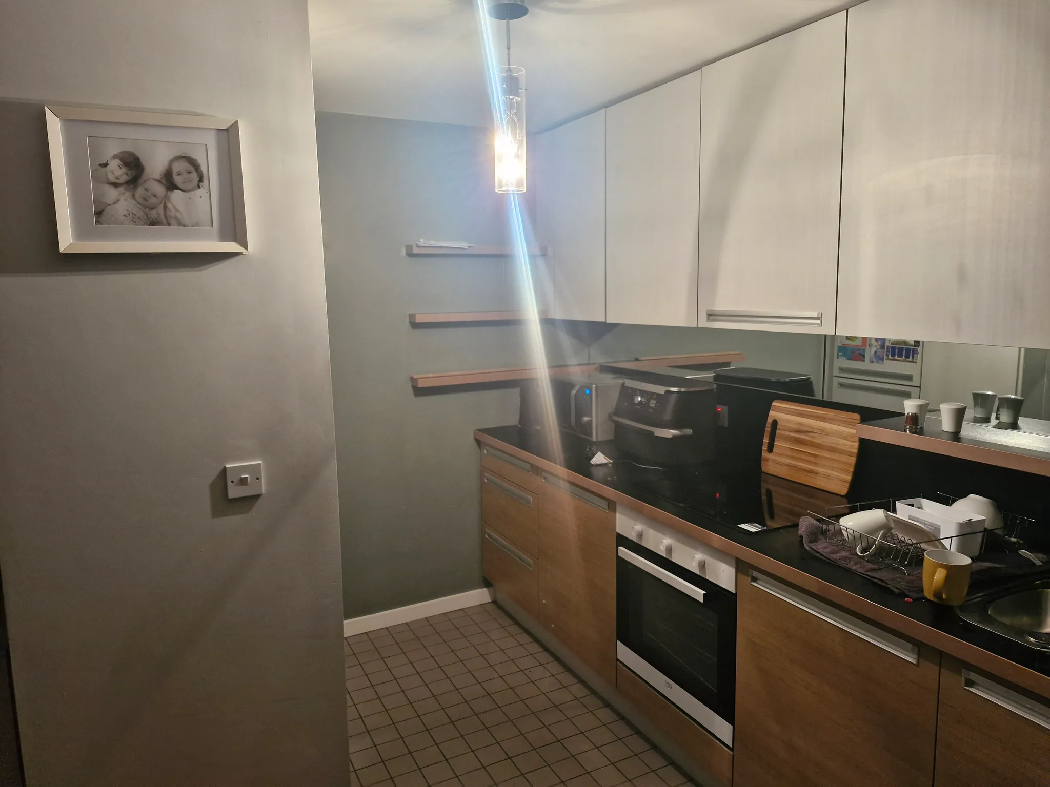 Small galley kitchen with wooden lower cabinets, white upper cabinets, black countertop, stovetop and sink, pendant light and a framed photo on the wall.