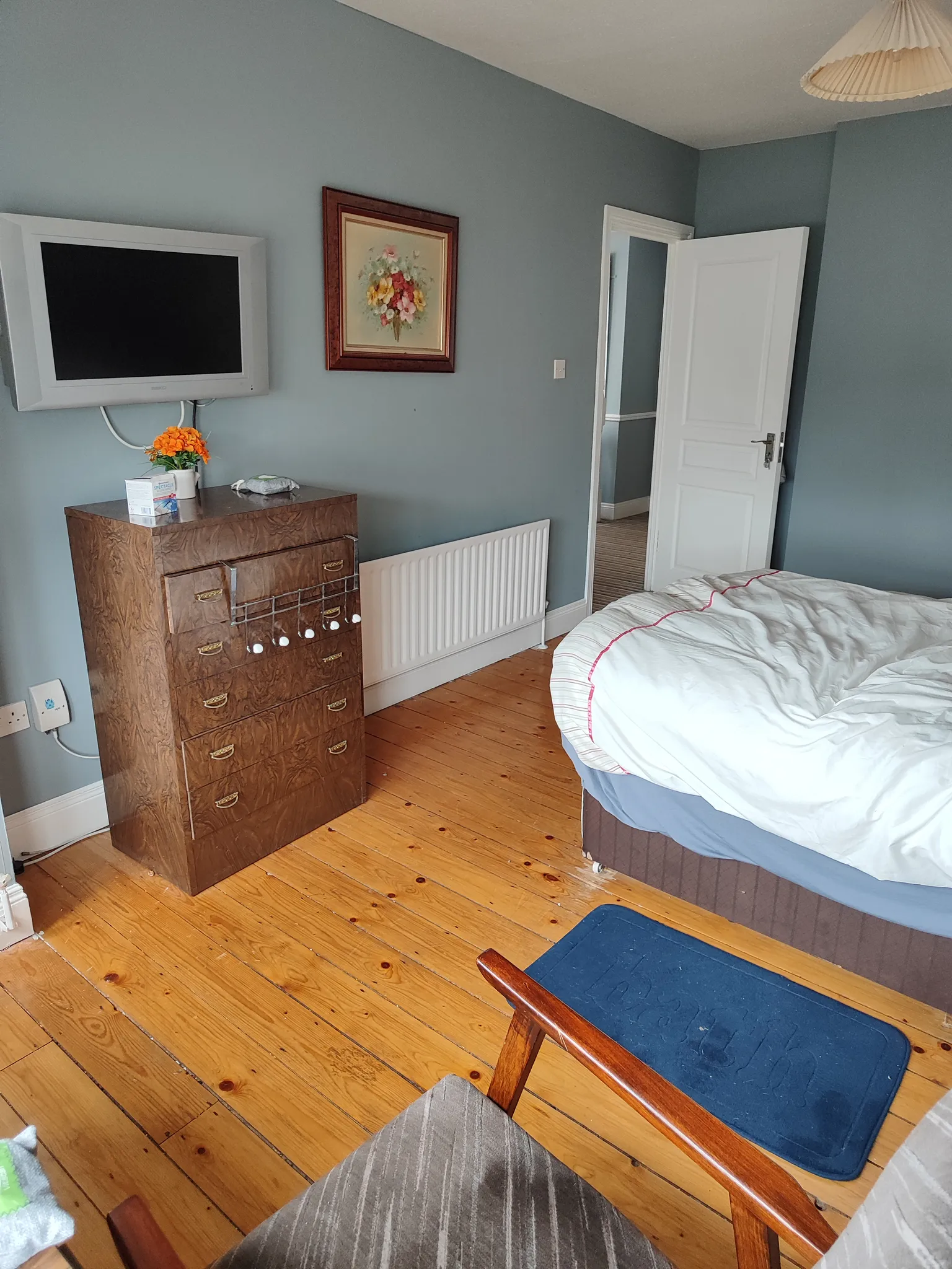 Bedroom with a bed, wooden dresser, wall-mounted TV, framed artwork, radiator and an open door.