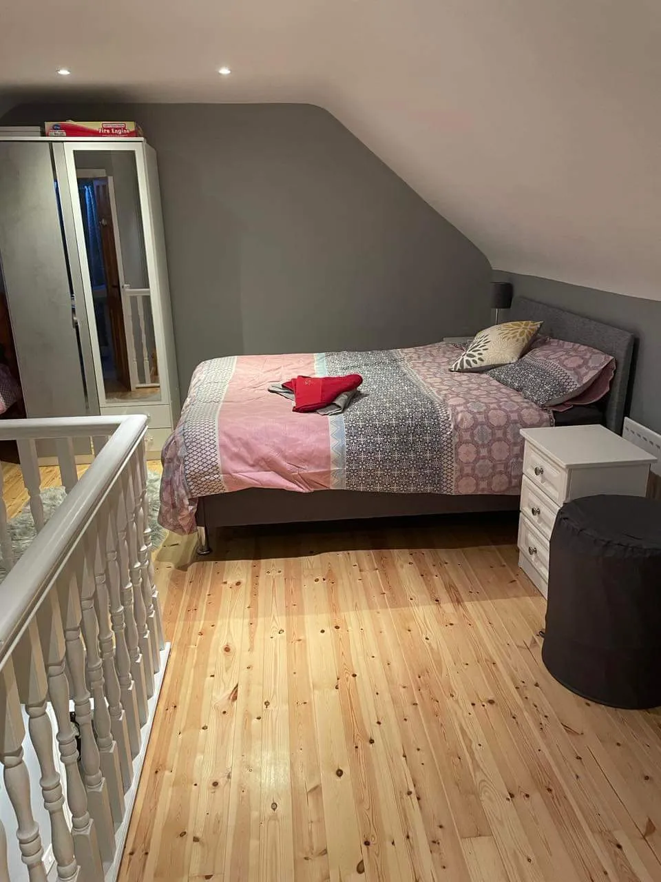 Bedroom with a double bed against a sloped ceiling, bedside table, wardrobe with mirrored door, wooden floor and railing in the foreground.
