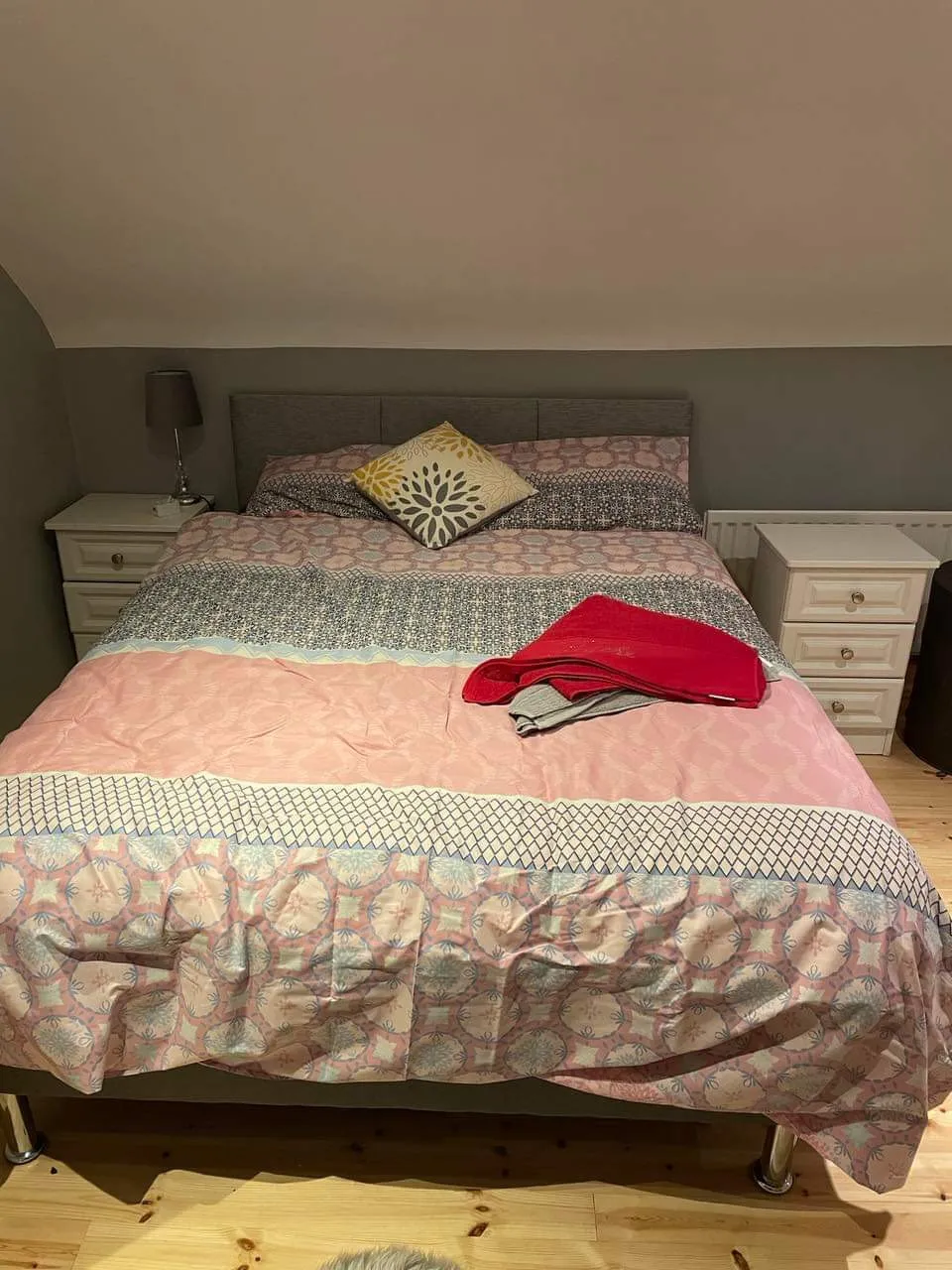 Bedroom with a double bed centered against a wall under a sloped ceiling, patterned duvet, decorative pillow, folded blankets on the foot of the bed, two bedside tables and a lamp.