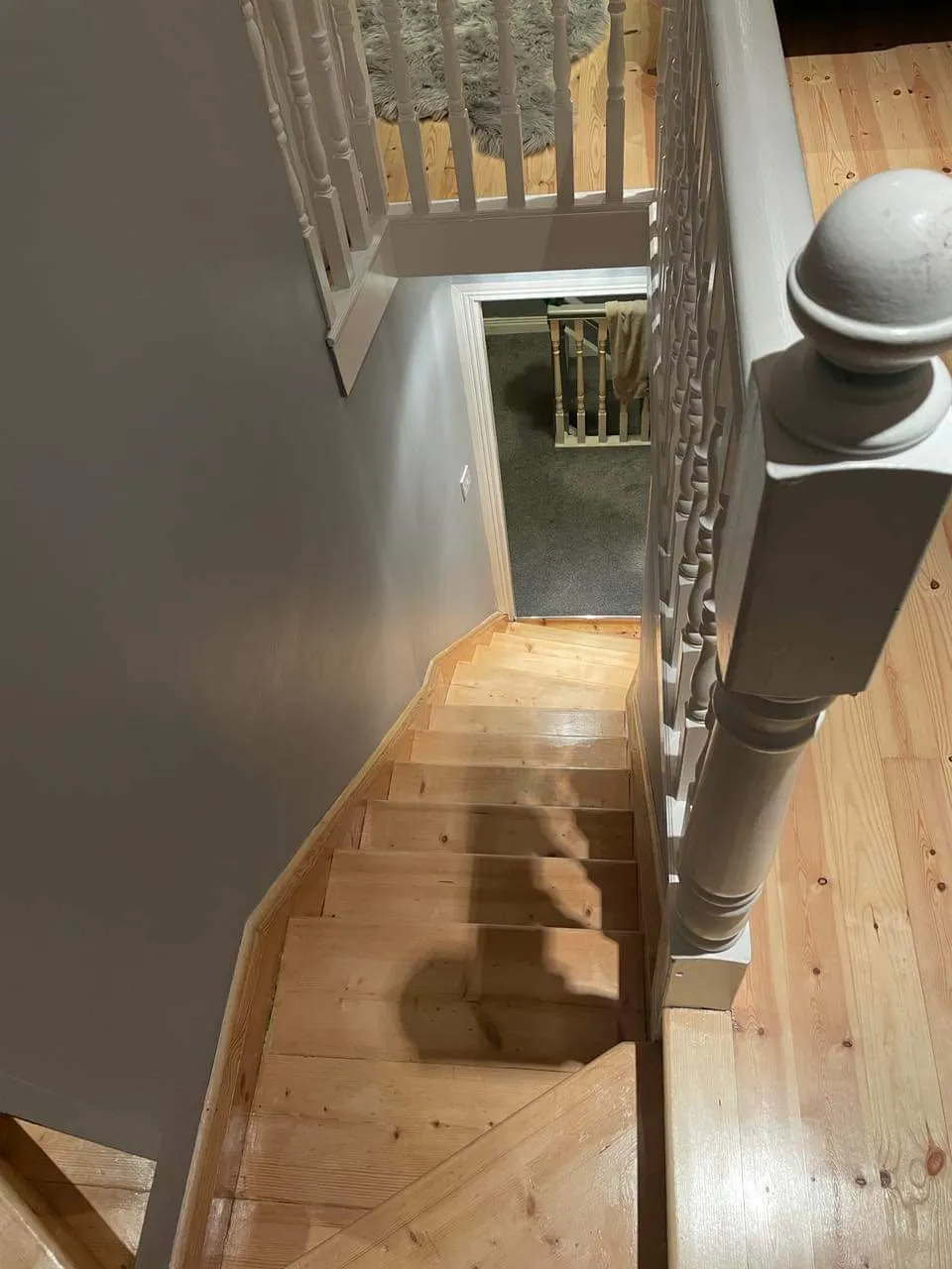 View down a wooden interior staircase with a white banister and a top landing leading to a doorway and carpeted lower room.