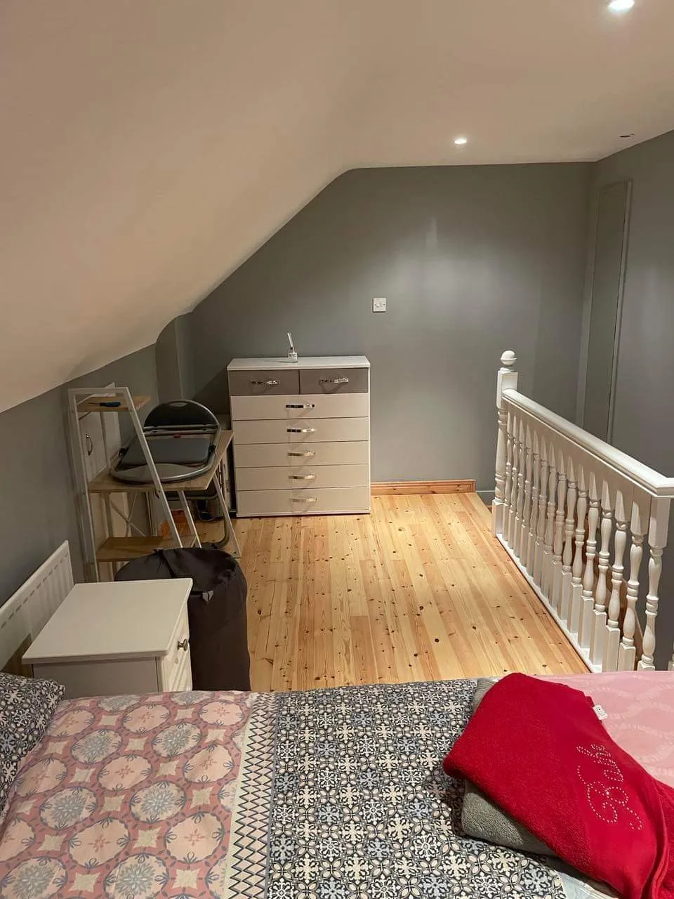 Bedroom loft area with a double bed in the foreground, wooden floor, white chest of drawers against a gray wall, and a white balustrade along the stair opening.