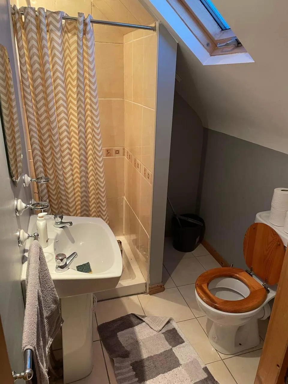 Bathroom with a shower and chevron curtain, pedestal sink with towel, toilet with wooden seat, tiled floor and a skylight