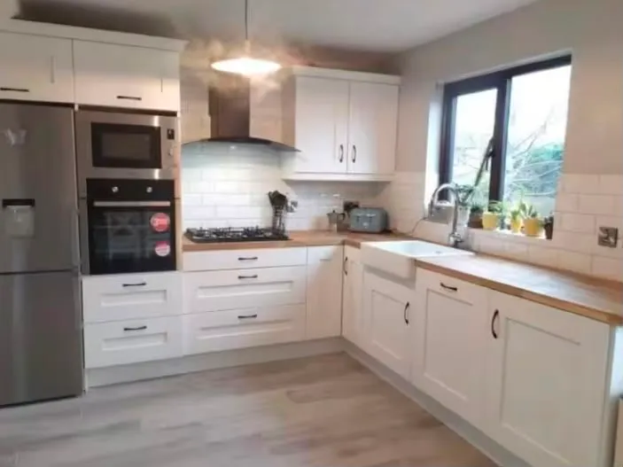Kitchen with white cabinets, wooden countertops, farmhouse sink under a window, gas cooktop with hood, built-in oven and stainless refrigerator.