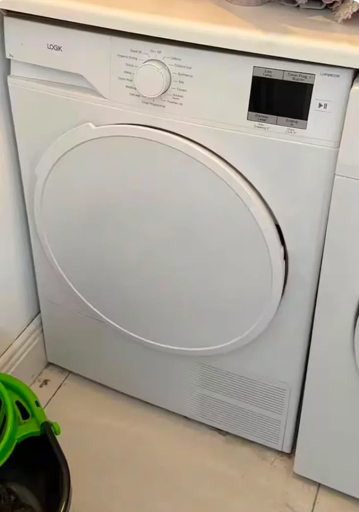 Front-loading white clothes dryer with round door, control knob and digital display, standing on a tiled floor.