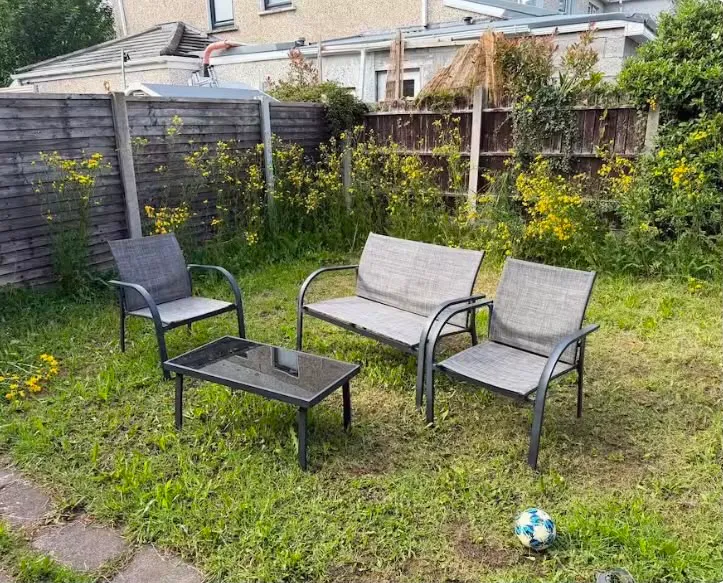 Backyard with a lawn, three outdoor seats (two chairs and a two-seat bench), a low glass-top coffee table, wooden fence, yellow wildflowers, and a soccer ball on the grass.
