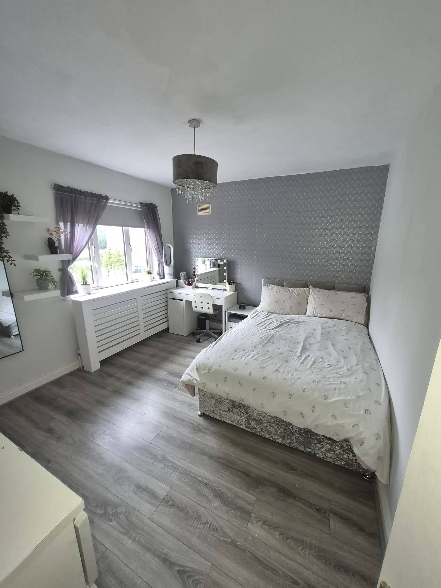 Bedroom with a double bed, patterned accent wall, bedside table, dressing table with mirror and chair, window with purple curtains and potted plants, and a ceiling light fixture.