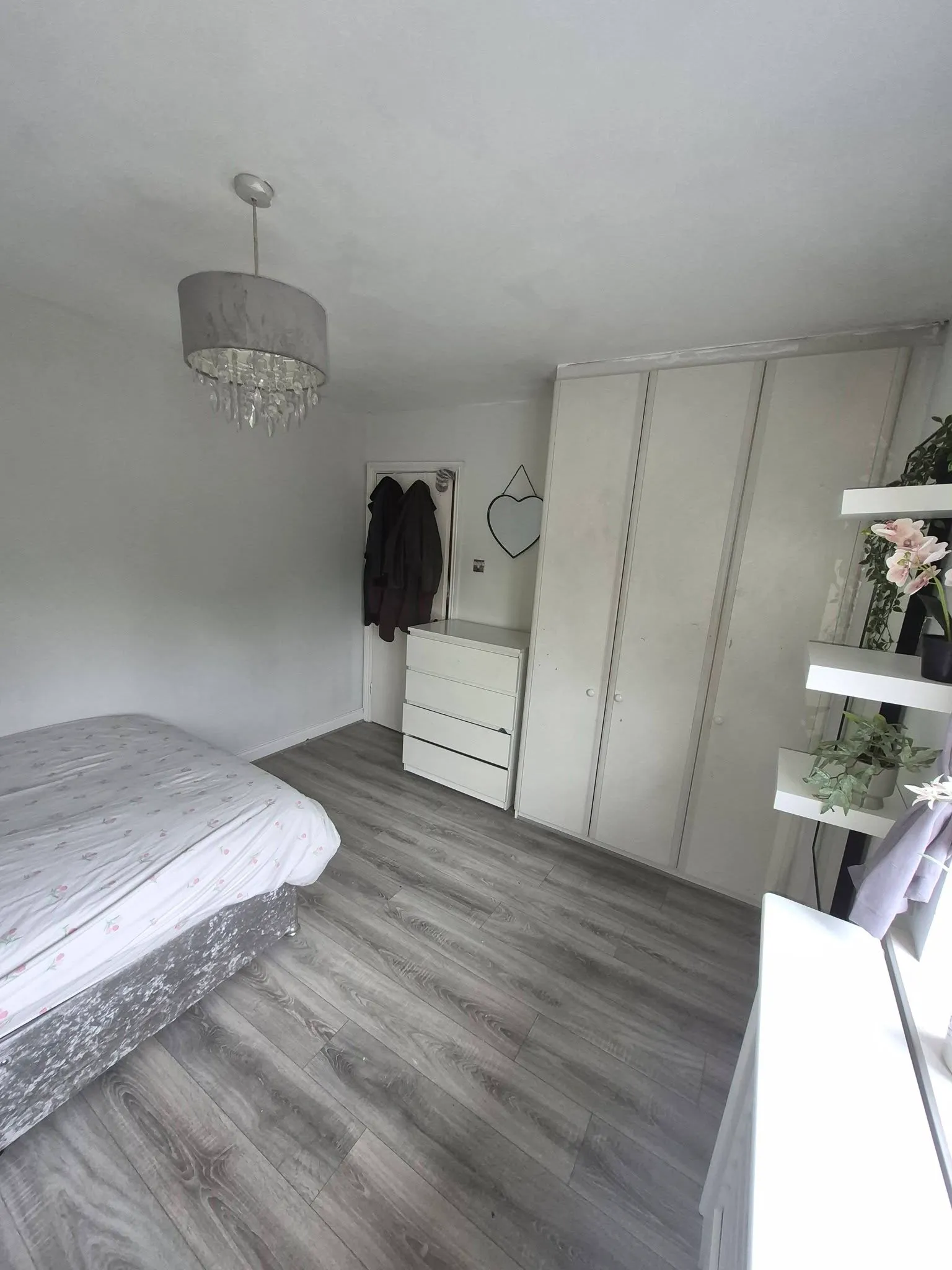 Bedroom with a visible double bed edge, a pendant light, tall wardrobe and chest of drawers, wall mirror in a heart shape, and shelving with plants near a window.