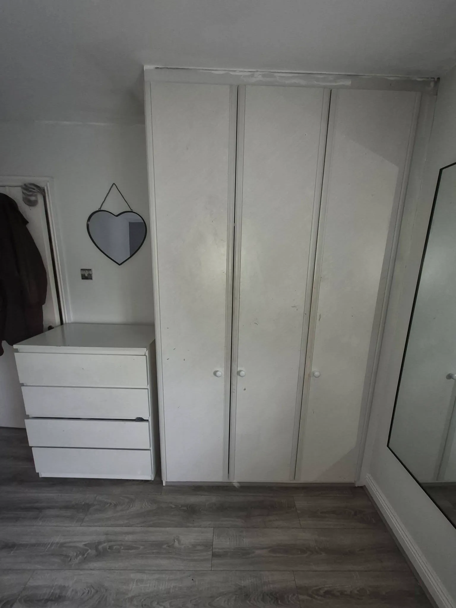 Bedroom corner with a tall wardrobe, a chest of drawers, a heart-shaped wall mirror and a floor mirror partially visible