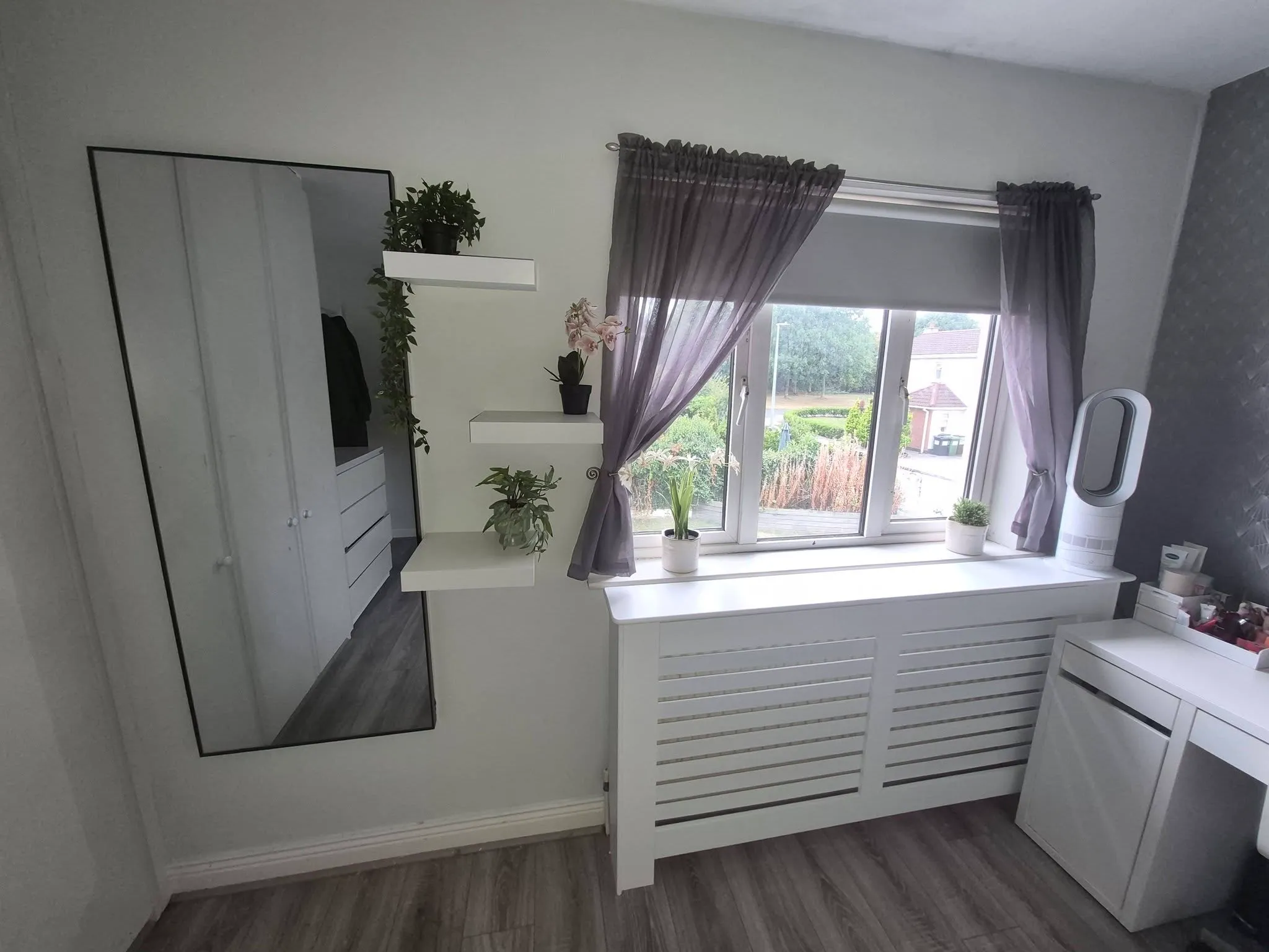 Bedroom corner with a large window with sheer curtains and roller blind, white radiator cover, wall-mounted shelves with plants, mirror and dresser.