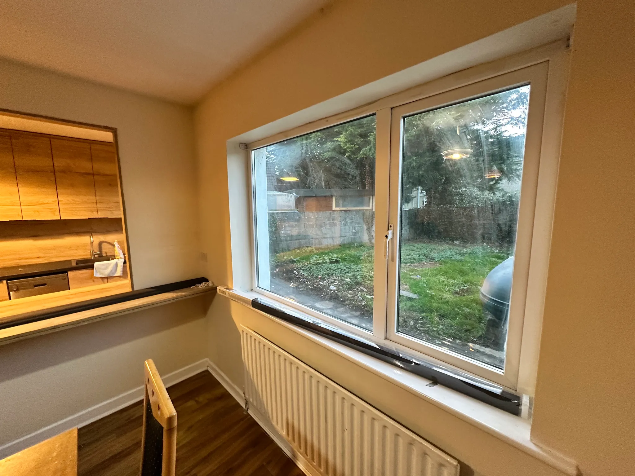 Dining area with a table and chair by a large window and radiator, view into a kitchen through a pass-through opening.