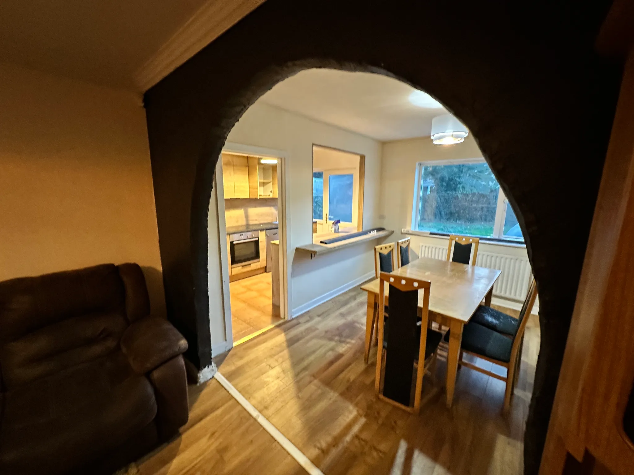 Dining room with a wooden table and chairs seen through a dark arched opening; adjoining kitchen with oven and a window visible.