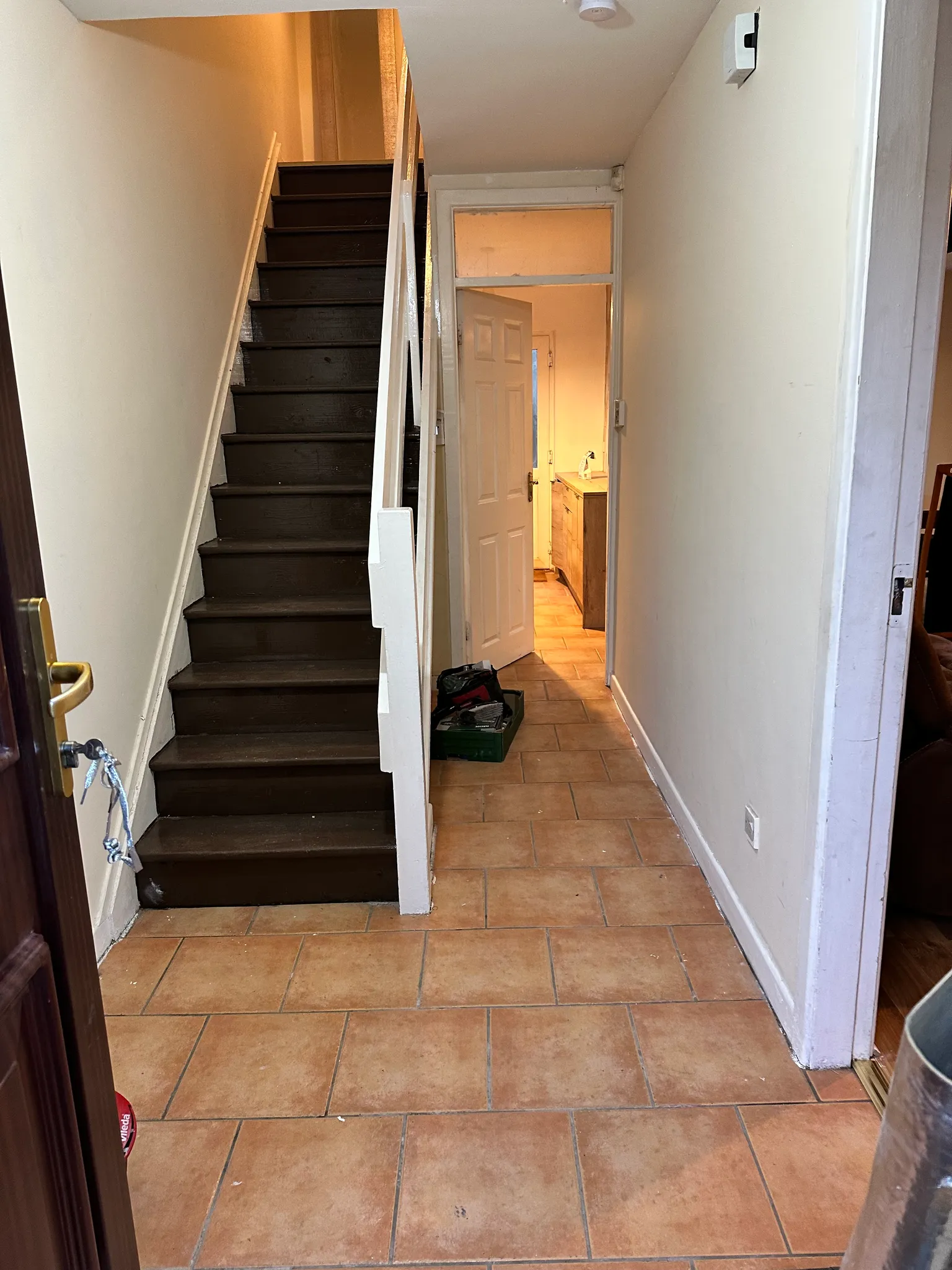Narrow interior hallway with tiled floor, dark wooden stairs and white handrail on the left, an open door ahead showing a countertop, and a small toolbox on the floor.