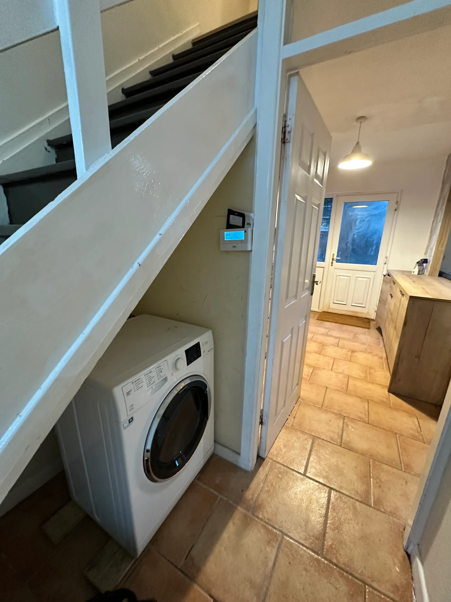 Hallway with stairs and a front-loading washing machine under the stairs, tiled floor, open interior door and back door with pendant light.