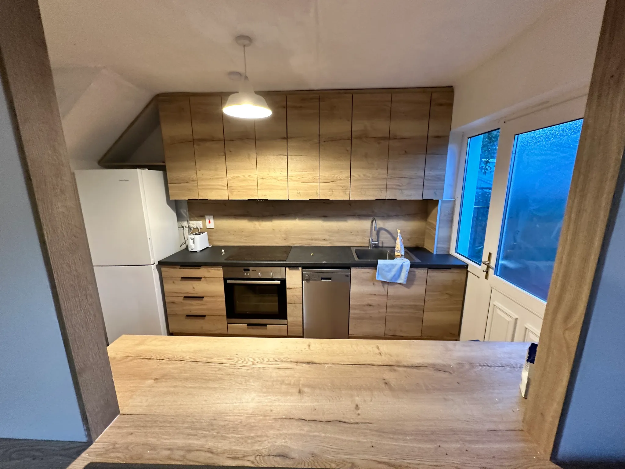 Kitchen with wood cabinets, black countertop, built-in oven, dishwasher, refrigerator, sink, pendant light and a door with frosted glass window.