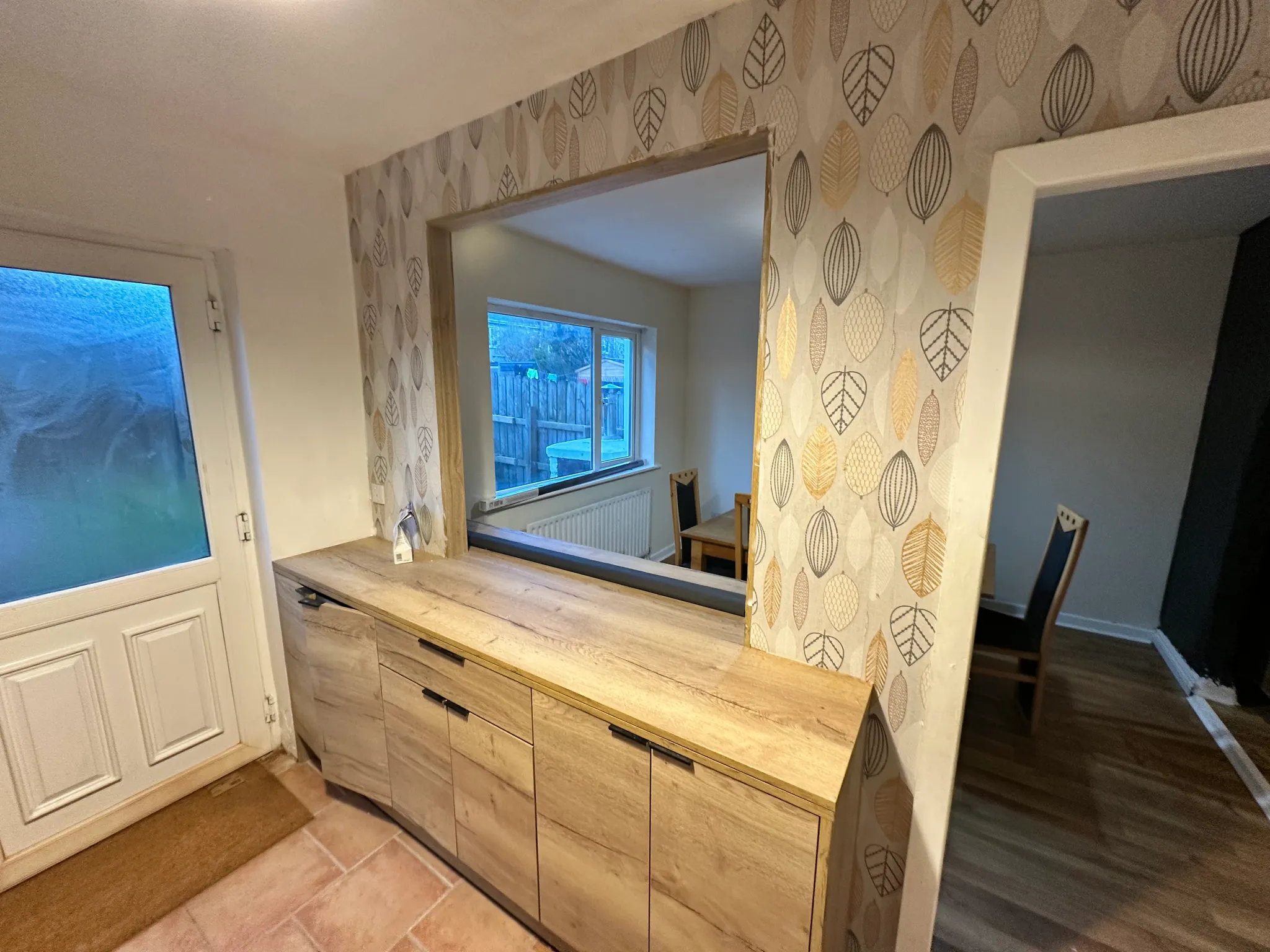 Kitchen area with wooden base cabinets and a pass-through opening to a dining area; patterned leaf wallpaper around the opening, a frosted-glass door at left and a window into the adjacent room.