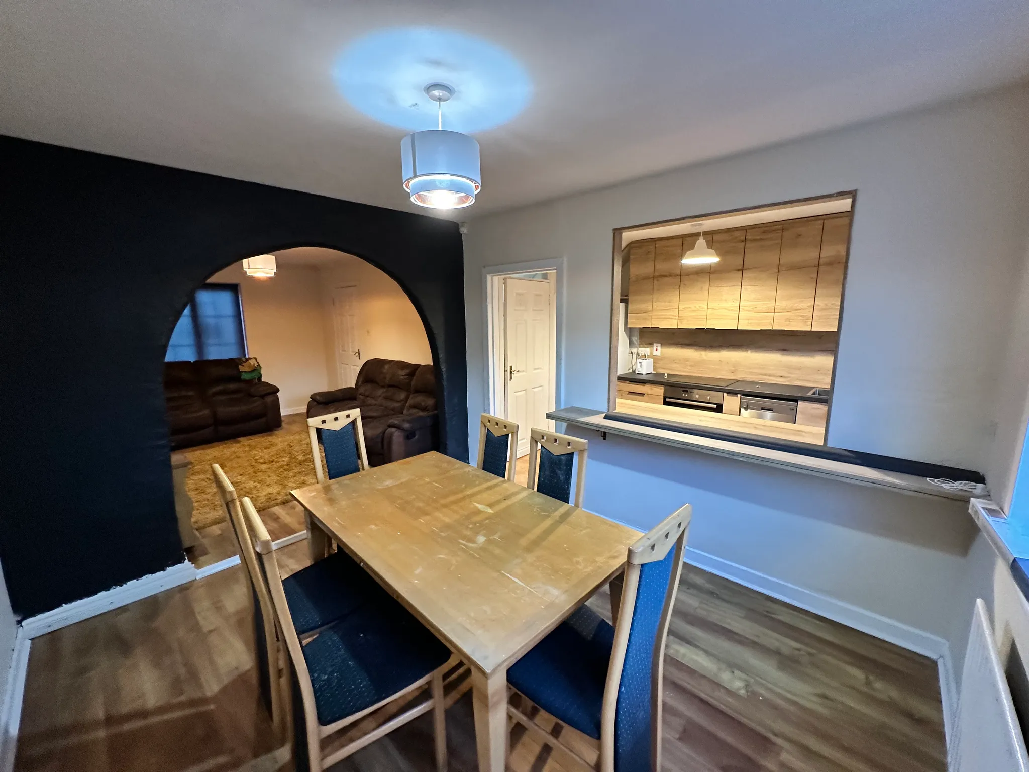 Dining room with a wooden table and six chairs, pendant light, open pass-through window to a kitchen with wooden cabinets, and an arched opening to a living area with recliner sofas.