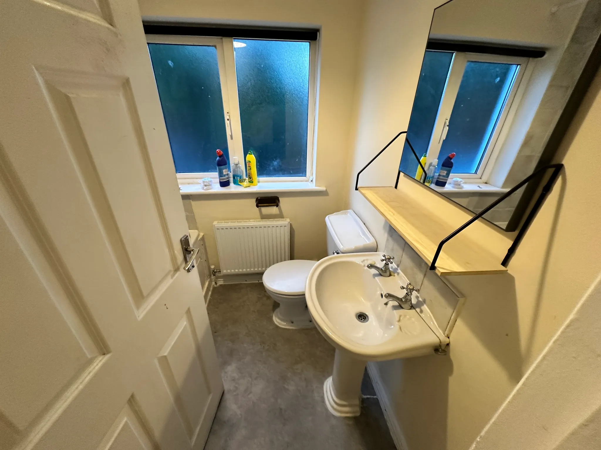 Small bathroom with a pedestal sink, toilet, frosted window, wall-mounted mirror with wooden shelf, radiator and cleaning bottles on the windowsill.